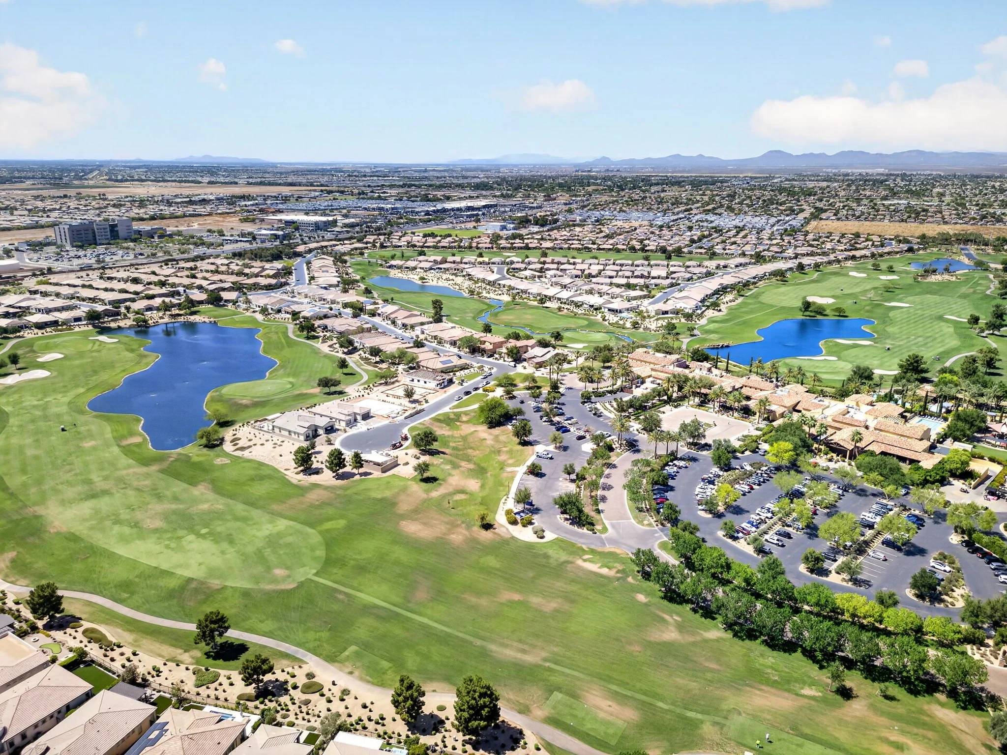 Aerial view of a suburban golf course with lakes, houses, and a parking lot, under a partly cloudy sky with mountains in the distance.