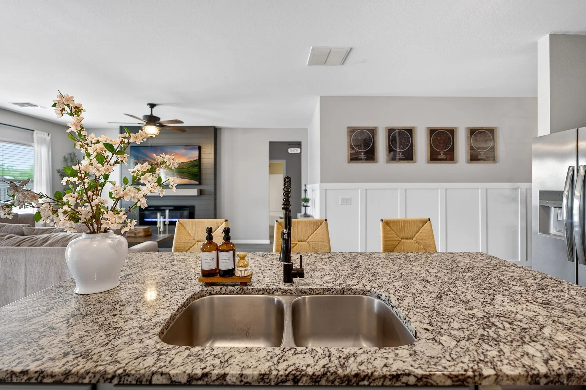 A kitchen island with a granite countertop, a vase with pink flowers, and bottles of soap in the foreground. In the background, there are yellow chairs, a ceiling fan, a television, and framed star charts on the wall.