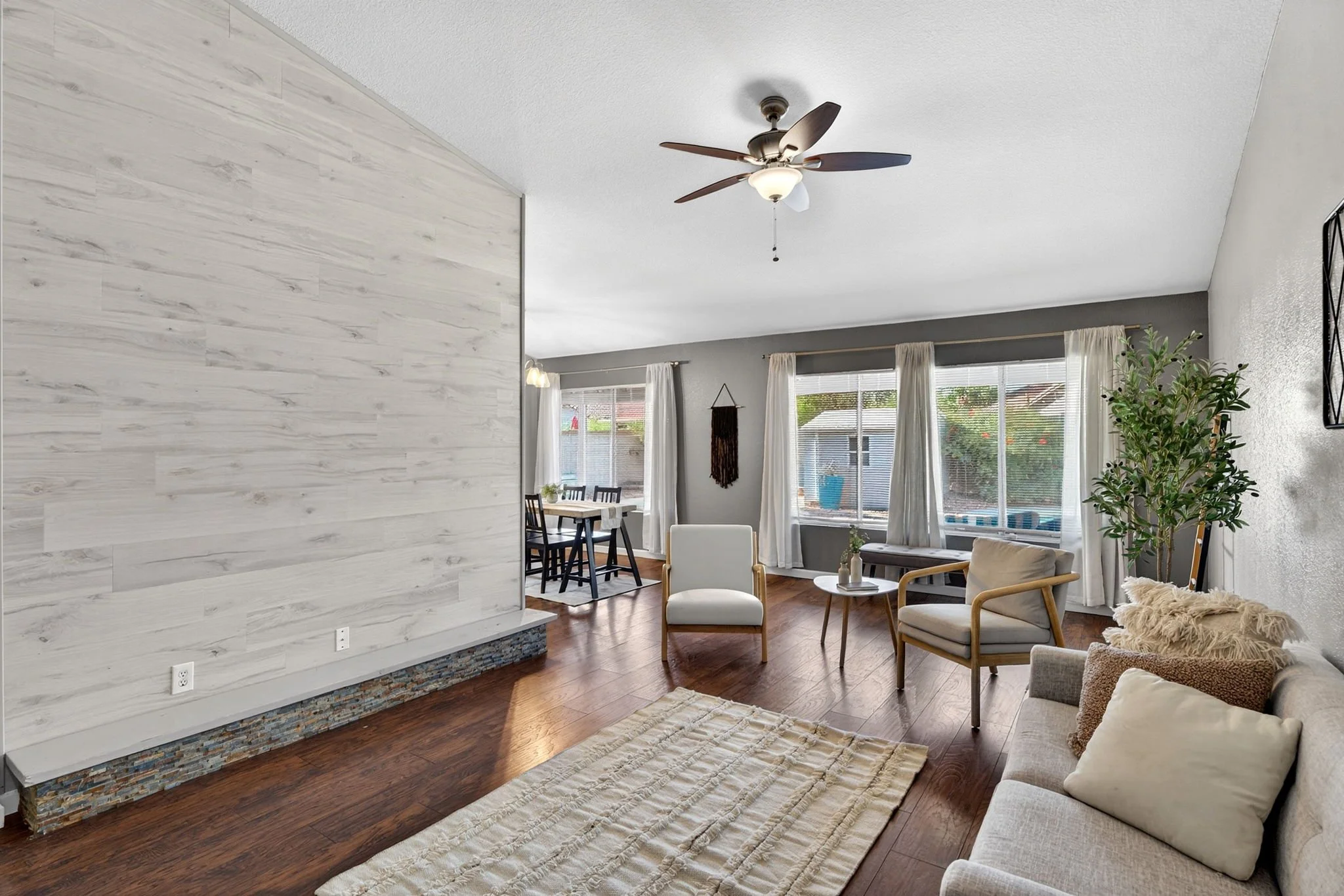 Living room with white walls, hardwood flooring, large windows, white curtains, a white and beige sofa with pillows, a wooden armchair, a small coffee table, a ceiling fan, a textured wall, and a view of the backyard with a pool and garden.