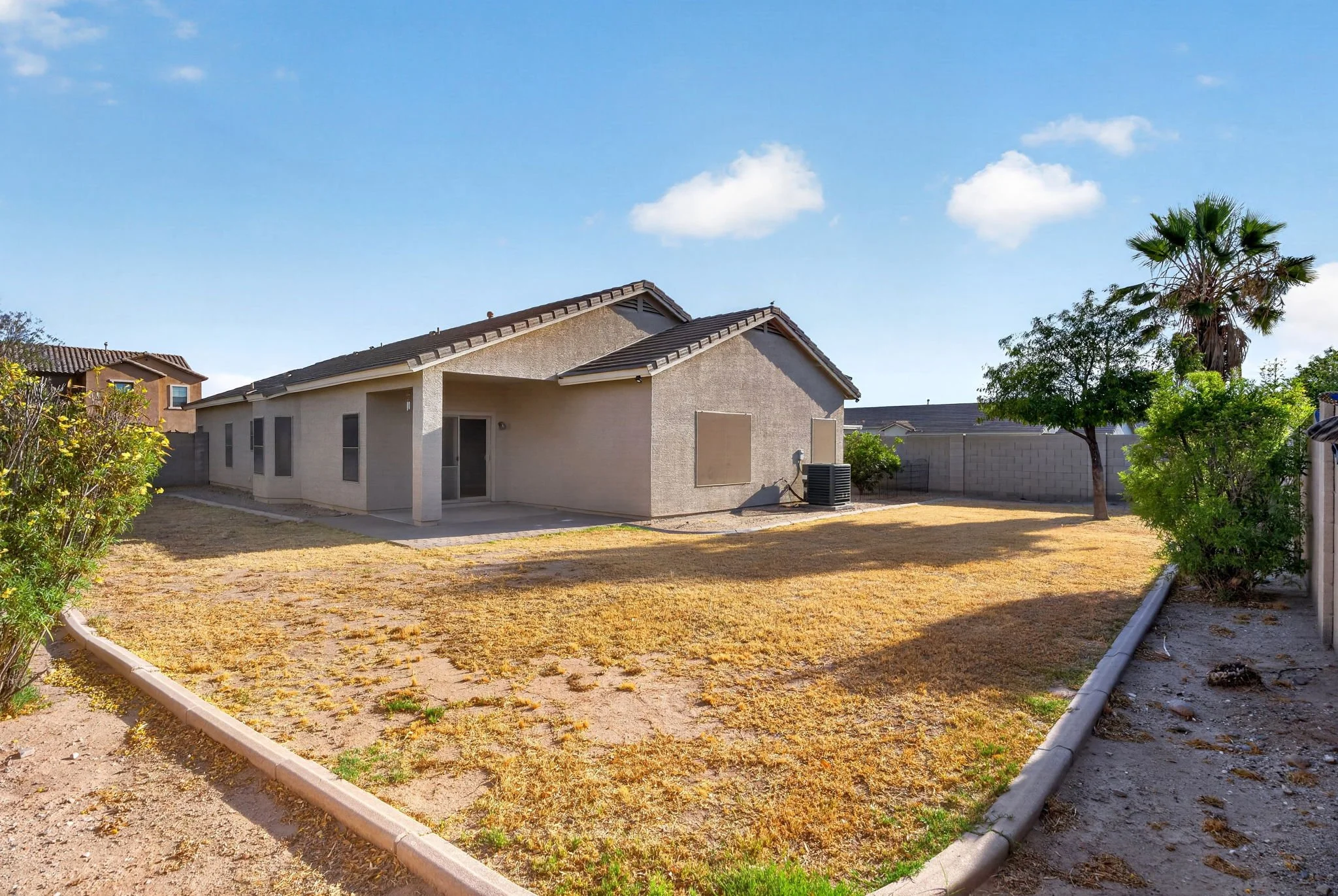 A backyard with a single-story house with beige stucco walls and a dark shingled roof. The yard has a mix of dirt and yellowed grass, surrounded by bushes, trees, and a concrete block wall. A small paved patio is adjacent to the house, with an air co