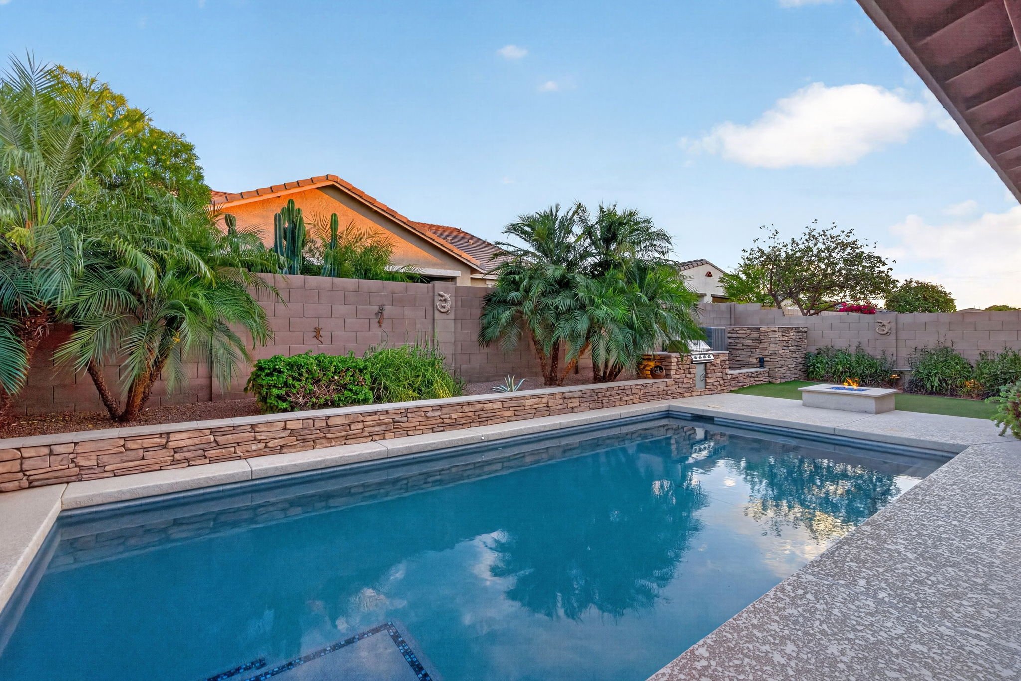 Backyard with swimming pool, lush green plants, palm trees, a fire feature, and a brick wall, under a clear blue sky.