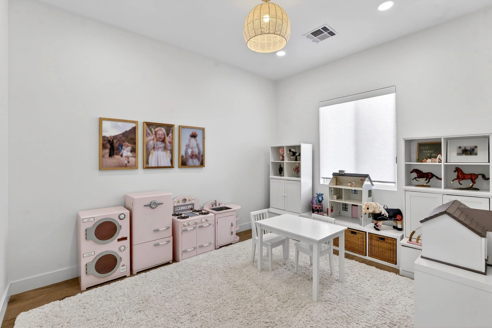 A children's playroom with white walls, a shaggy white rug, and natural light. There are three framed photos of children on the wall, a pink toy kitchen set, a small white table with chairs, and various toys and decorations, including horse figurines