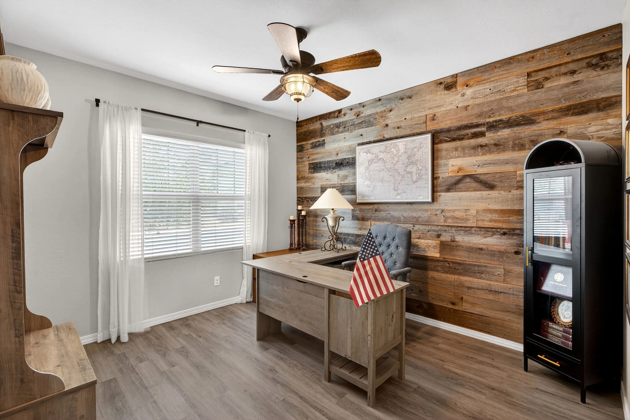 An office space with a wood accent wall, a desk with a chair, an American flag, a lamp, a world map on the wall, white curtains over a window, a ceiling fan, and a black display cabinet.
