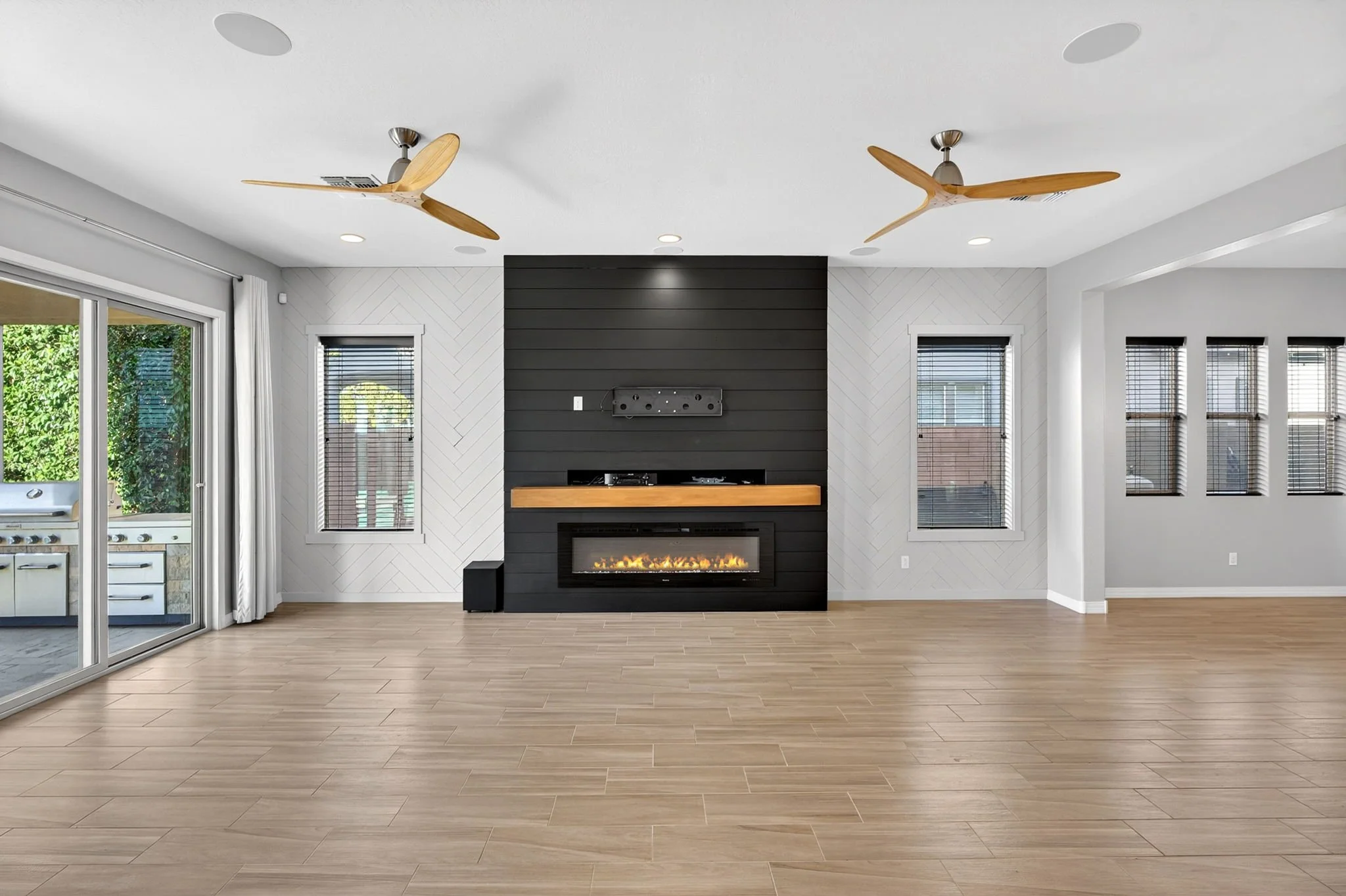 Interior of a modern living room with a fireplace, black accent wall, multiple windows with blinds, ceiling fans, hardwood flooring, and a sliding glass door leading outside.