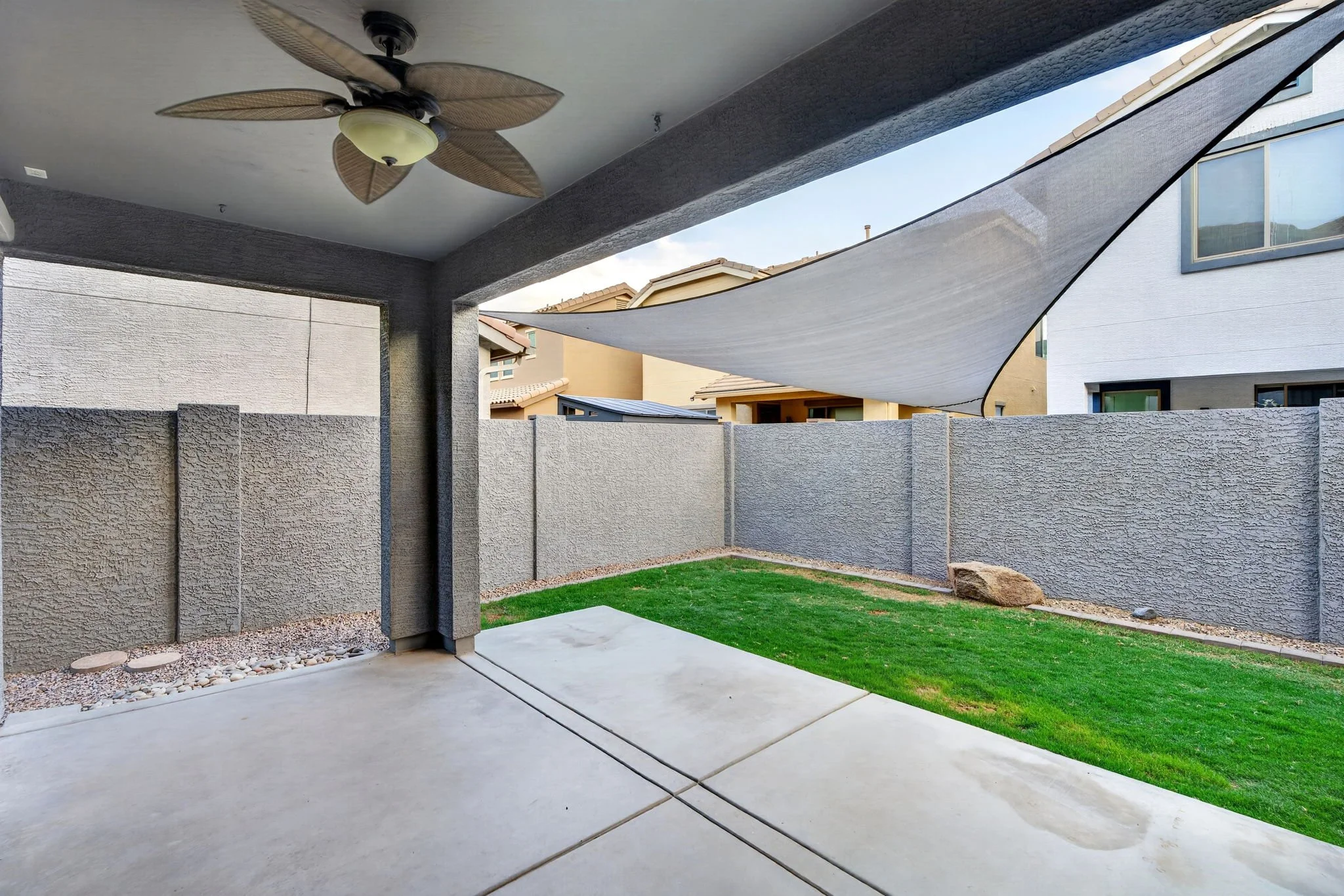 A covered patio area with a ceiling fan, a gray stucco wall, a small lawn, and neighboring houses visible beyond a gray fence, with a sunshade fabric stretched across part of the patio.