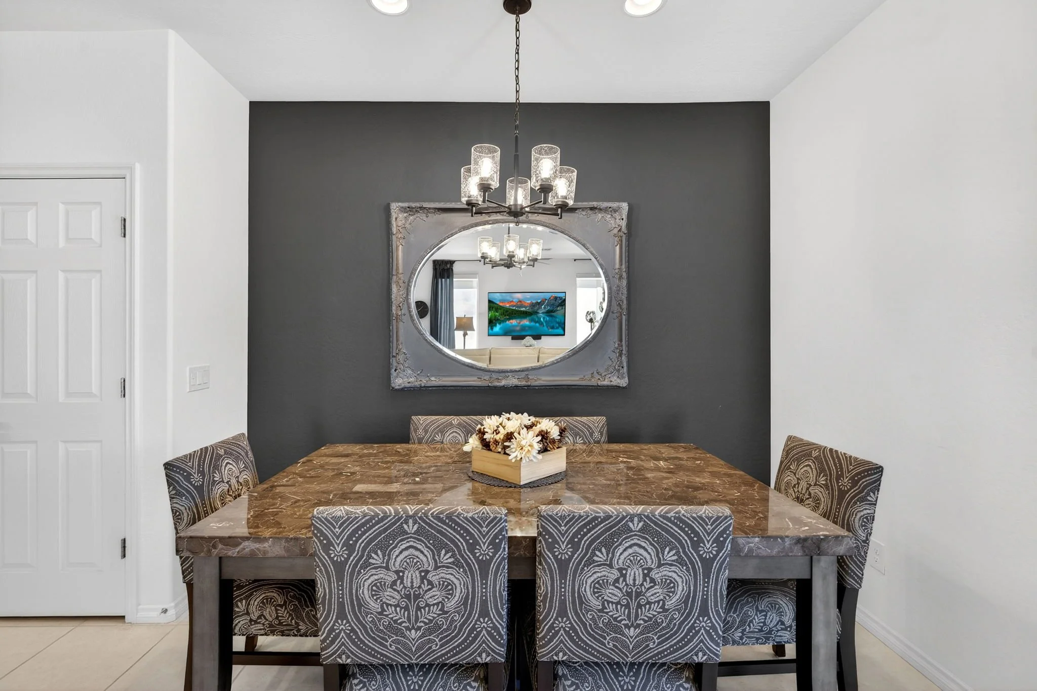 A dining room with a wooden table, six patterned chairs, a dark accent wall, a mirror, and a chandelier.