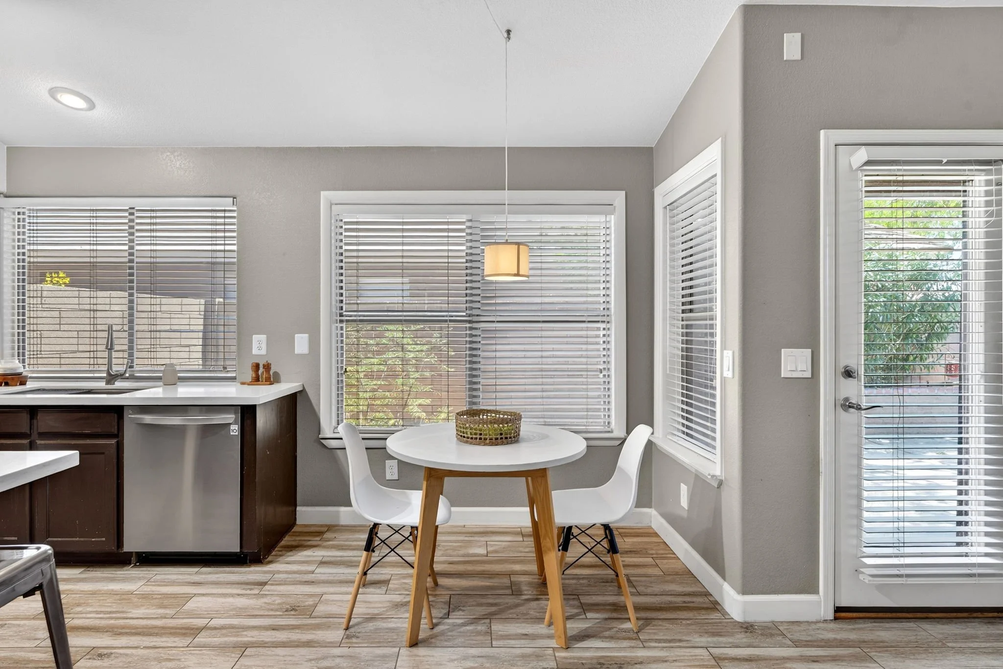 A kitchen with a small round dining table and three white chairs, large windows with blinds, a hanging light, and a door with blinds leading outside. The kitchen has gray walls, a white countertop, and dark brown lower cabinets.