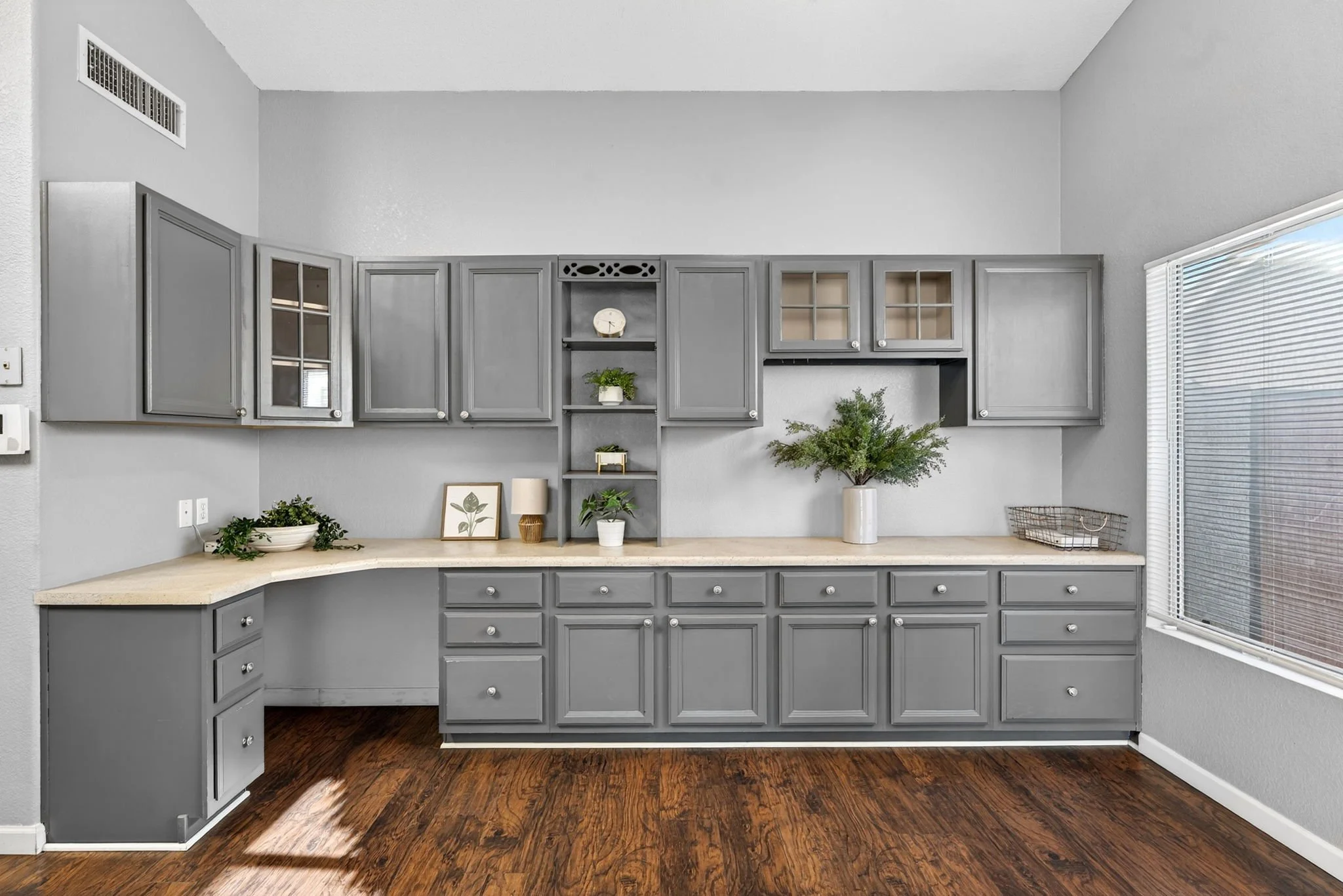 Gray kitchen cabinets on wall with a beige countertop, decorated with plants, framed pictures, a lamp, and a wire basket, under a window with blinds.