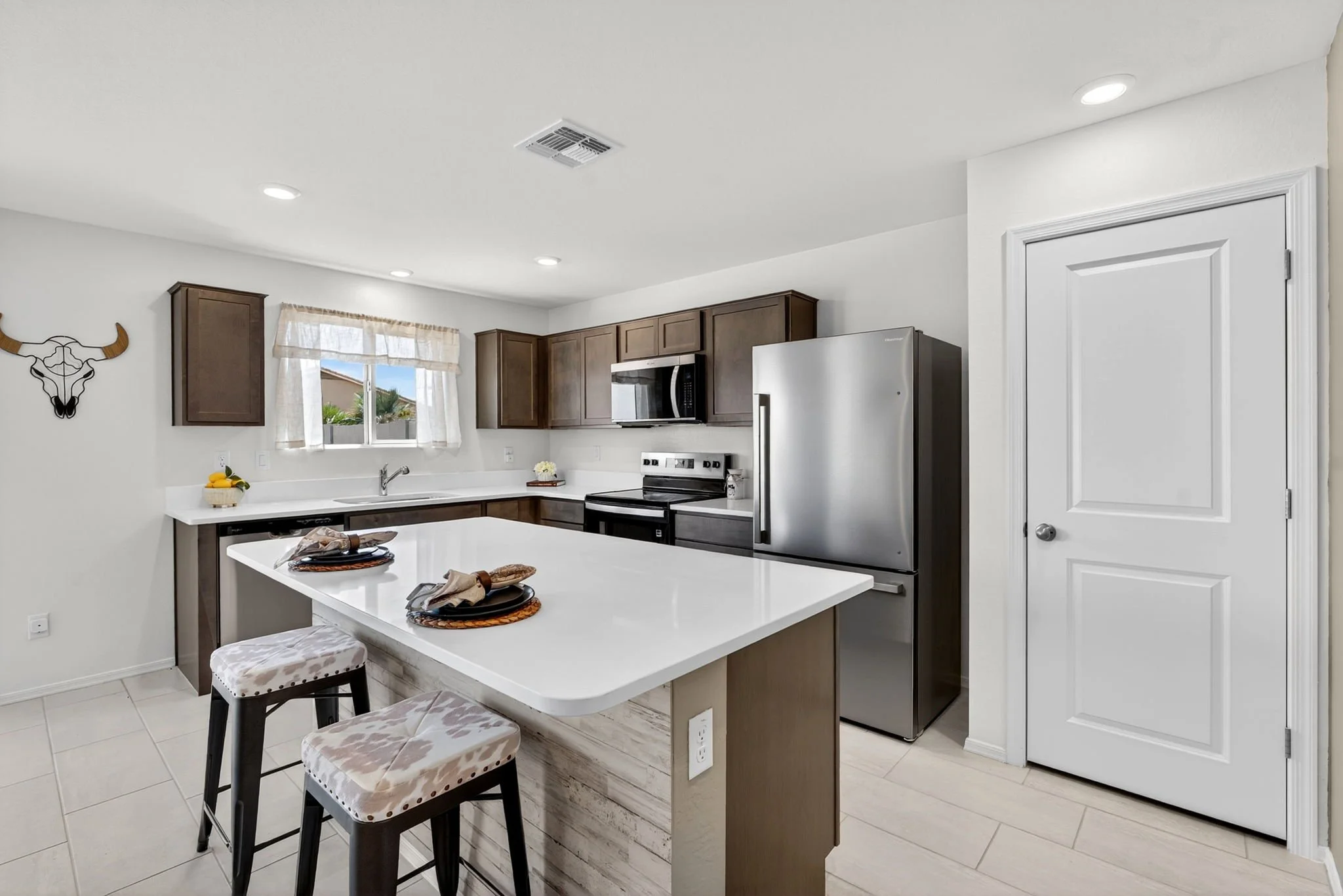 Modern kitchen with white countertops, dark wooden cabinets, stainless steel appliances, a window with curtains, and a white island with barstools.