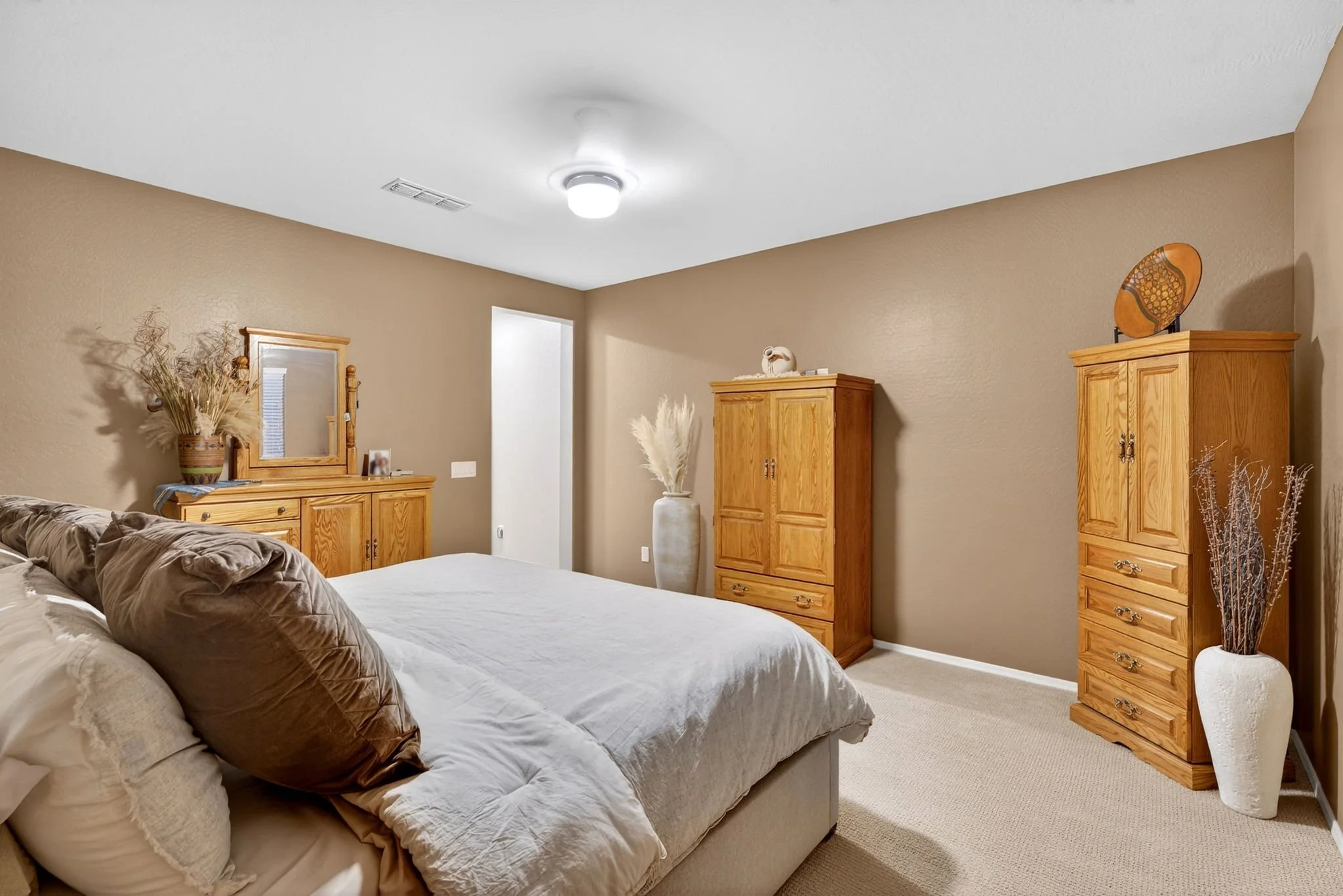 A bedroom with beige walls and carpet, featuring a large bed with white and brown bedding, wooden cabinets, decorative vases, and a ceiling light.