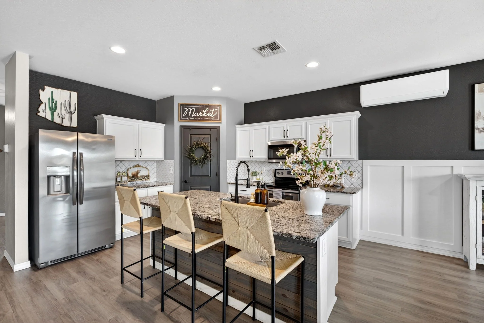 Modern kitchen with white cabinets, stainless steel refrigerator, granite countertop island, black wall, and decorative accents including a flower vase and wall art.