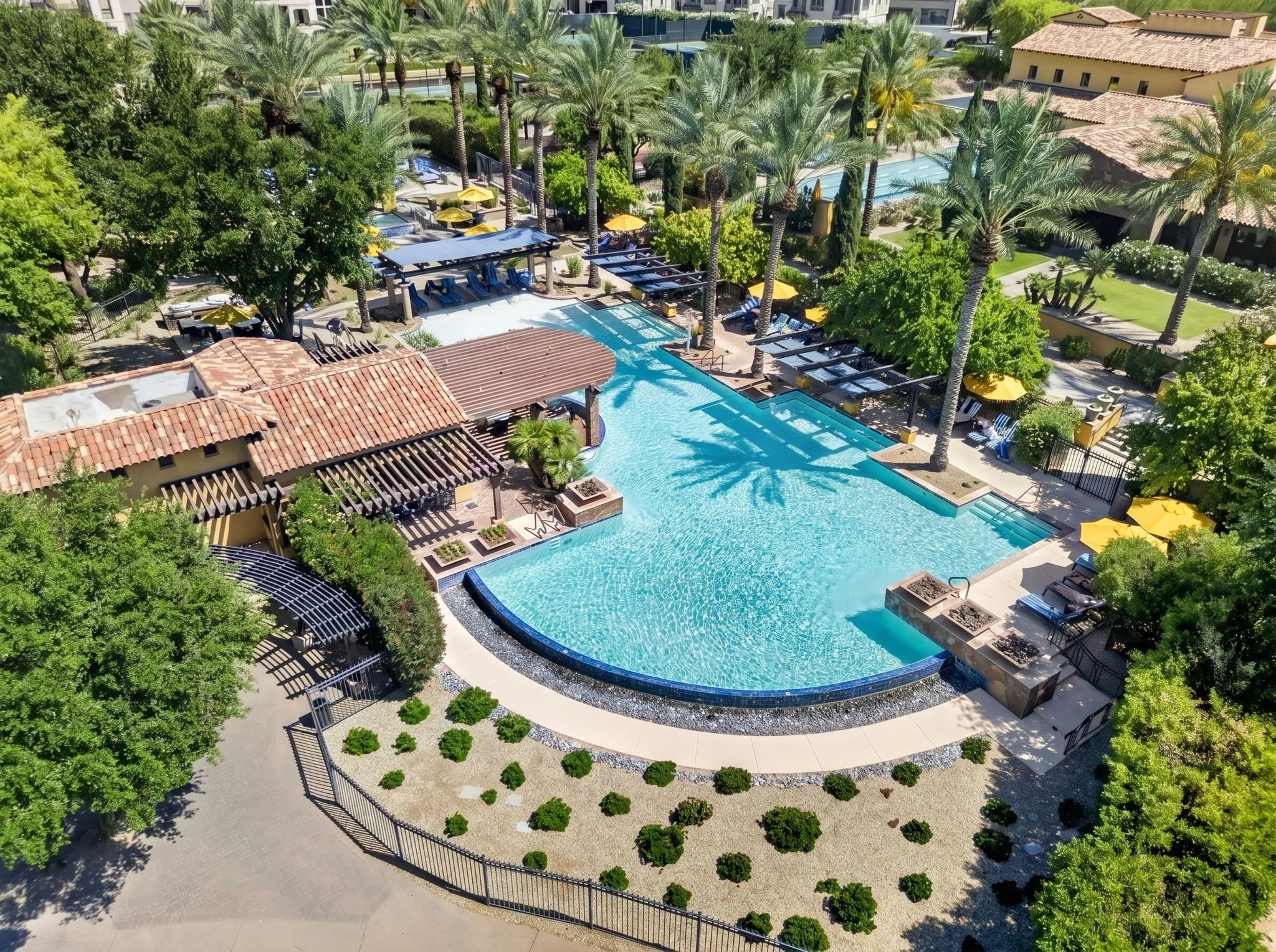 Aerial view of a luxurious outdoor swimming pool area surrounded by tall palm trees, sun loungers with blue cushions, yellow umbrellas, and shaded cabanas in a tropical resort setting.