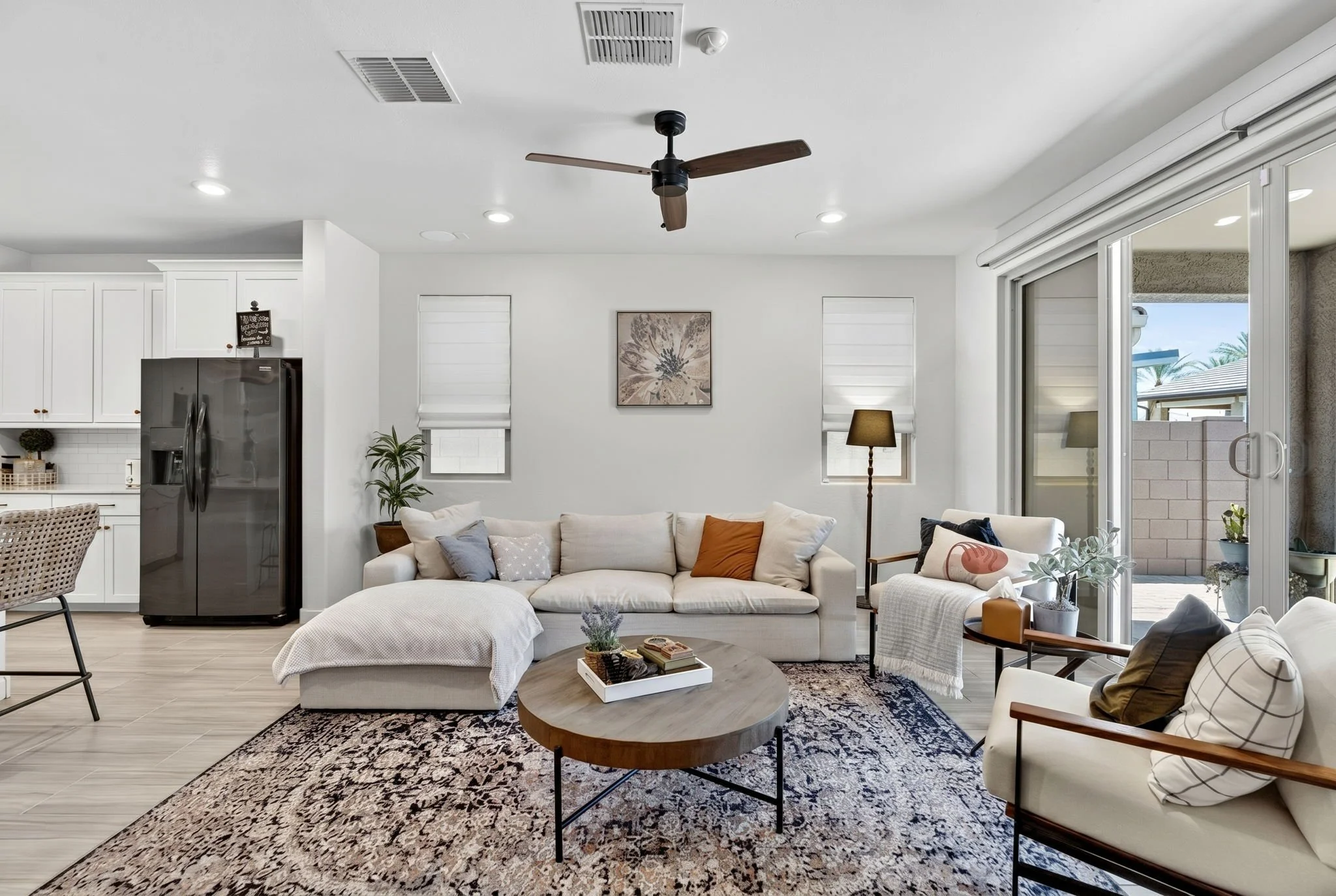 Living room with white sofa, coffee table, patterned rug, armchair, large window with blinds, sliding glass door, ceiling fan, ceiling lights, wall art, potted plants, and outdoor view of palm trees.