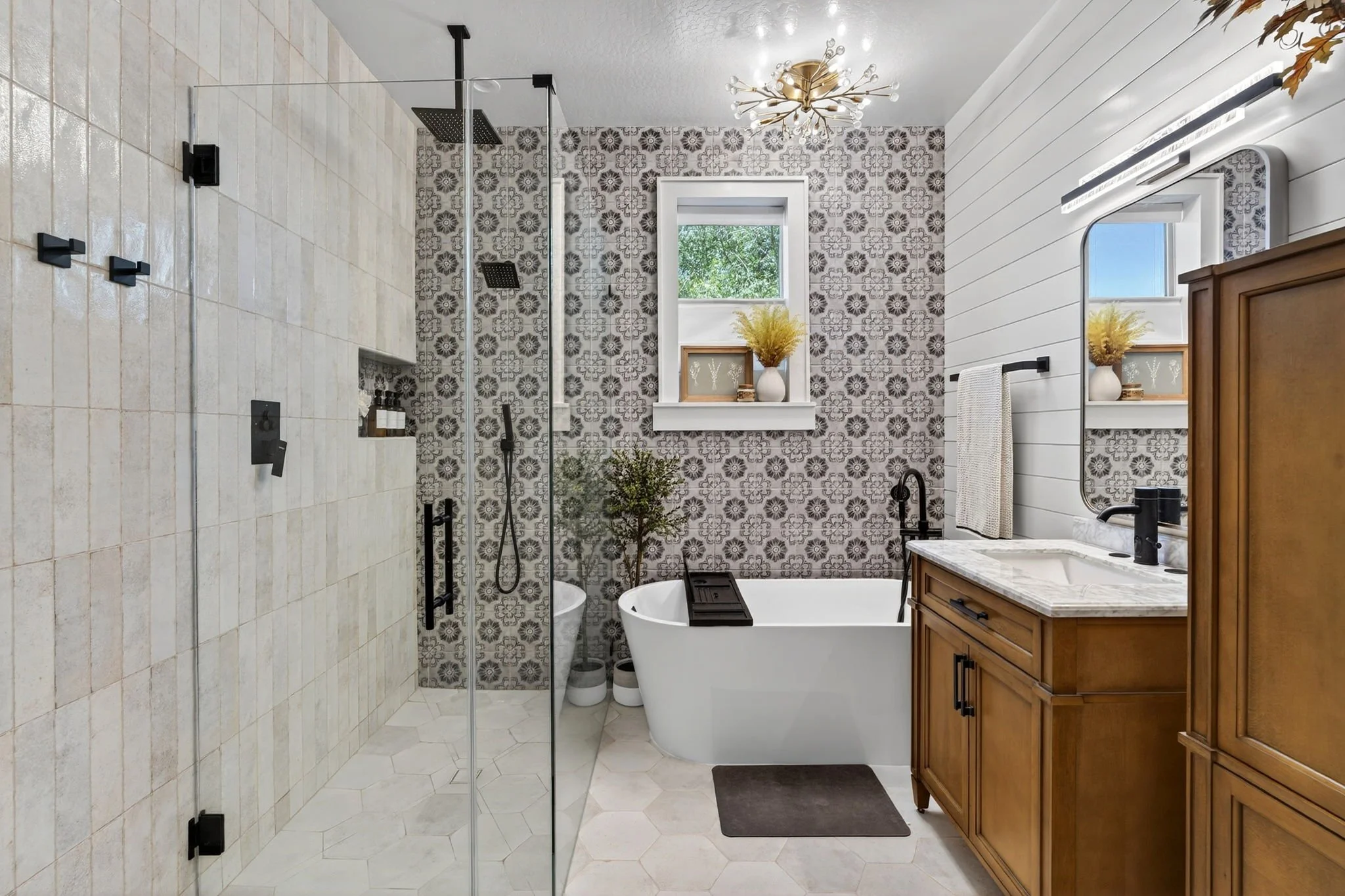 A bathroom with a walk-in shower on the left, a bathtub under a window in the center, and a wooden vanity with a mirror on the right. The wall behind the bathtub has patterned tiles, and the ceiling features a modern chandelier. There are decorative 