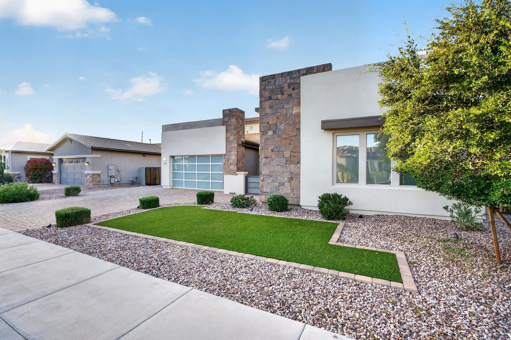 Modern house with a white exterior, stone accents, large window, and a garage. Front yard has manicured grass, small bushes, gravel, and walkway. A large tree is on the right side, and a neighboring house is visible in the background.