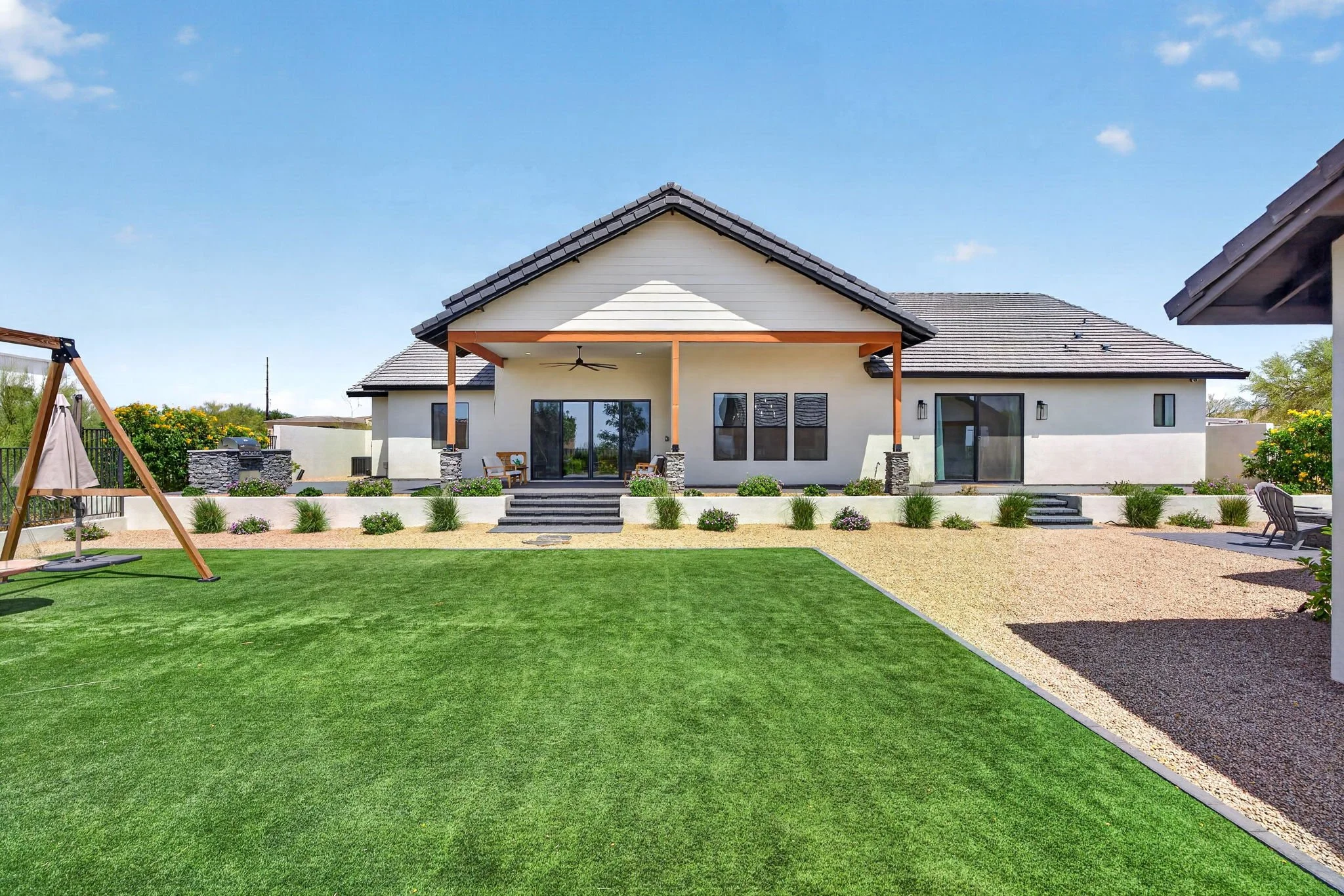 Backyard with a patio, green lawn, swing set, and a modern house with a covered porch and glass sliding doors.