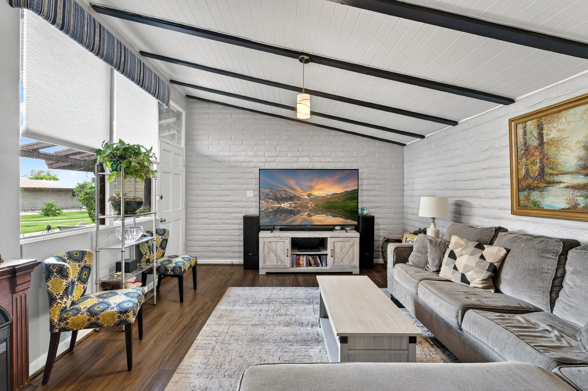 Living room with white brick walls, a gray sofa, a TV on a white stand, a painting, a window with blinds, a potted plant, and chairs with colorful patterns.