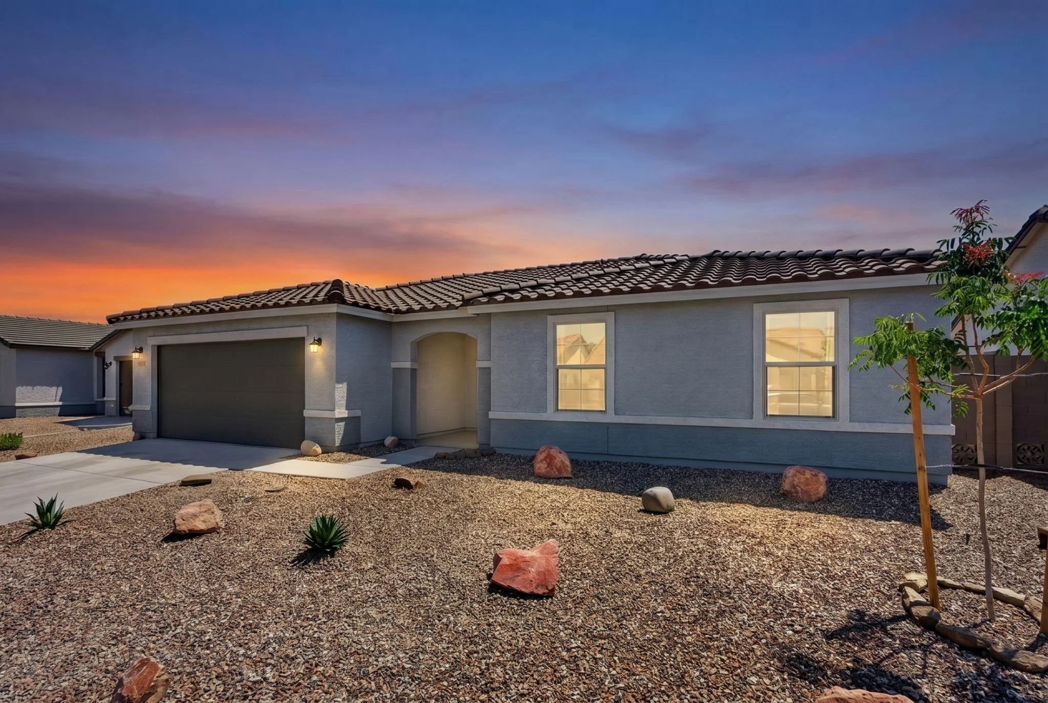 Front view of a single-story house during sunset with a gravel yard, small plants, a young tree, and a garage door, under a colorful sky.