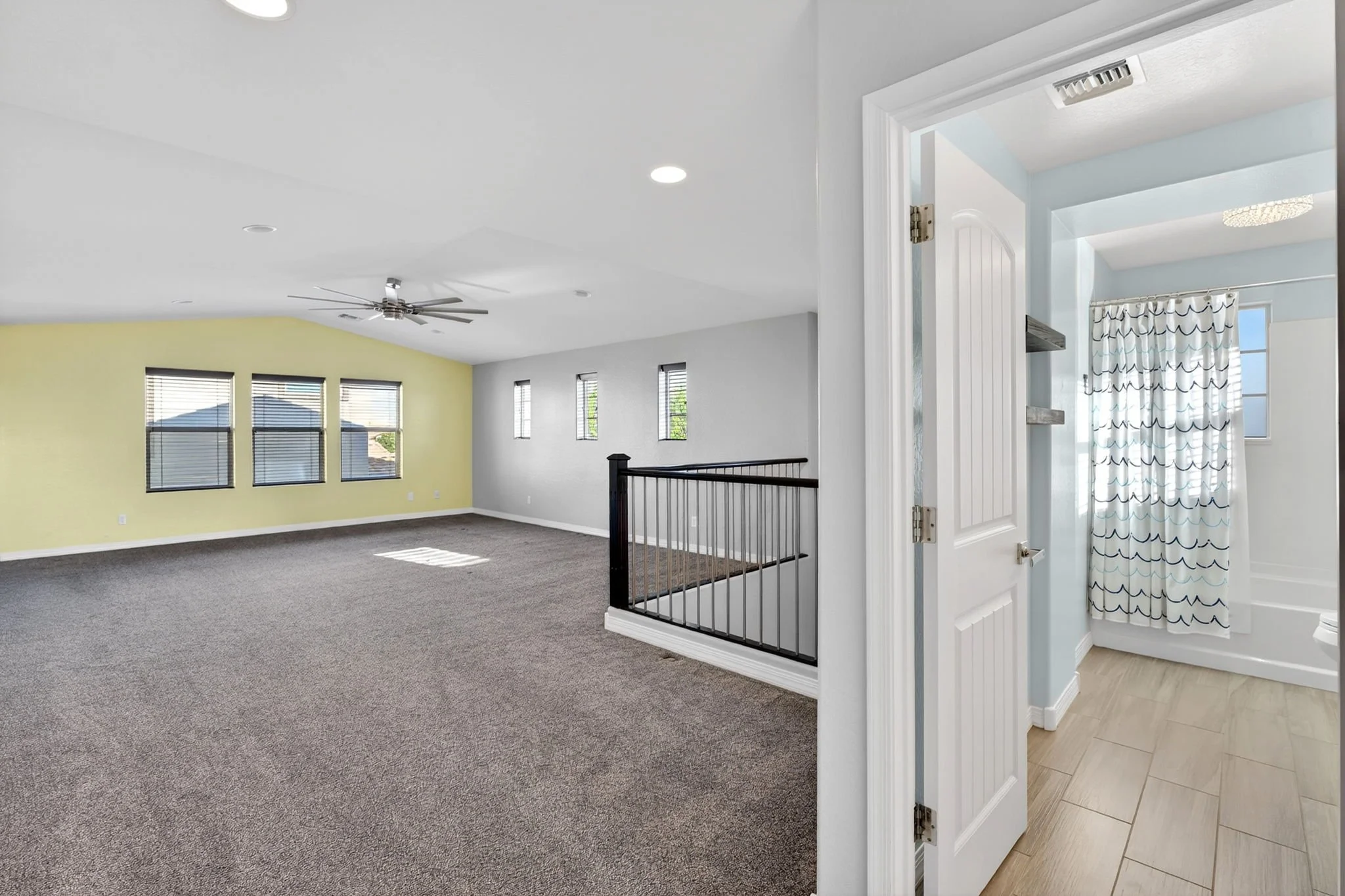 Interior view of a house showing a carpeted living area with a ceiling fan and multiple windows, and a doorway leading to a room with a shower curtain and tiled floor.