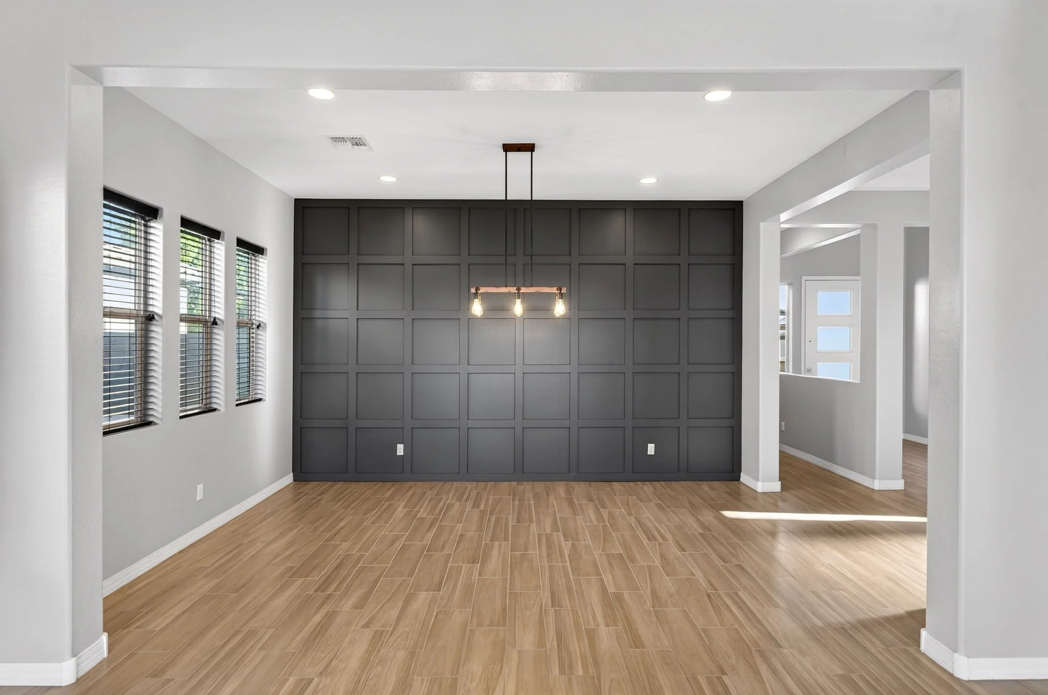 Interior view of a modern living space with a gray accent wall, wooden flooring, and large windows with black blinds, illuminated by ceiling and pendant lighting.