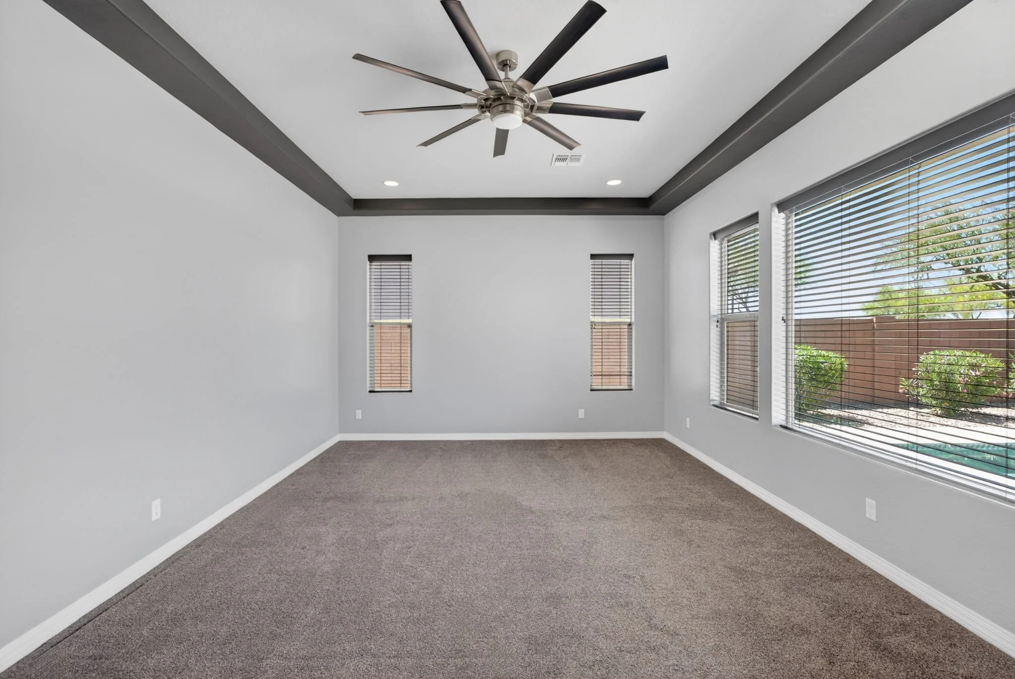 Empty room with gray walls, large windows with blinds, a ceiling fan, and carpet flooring.