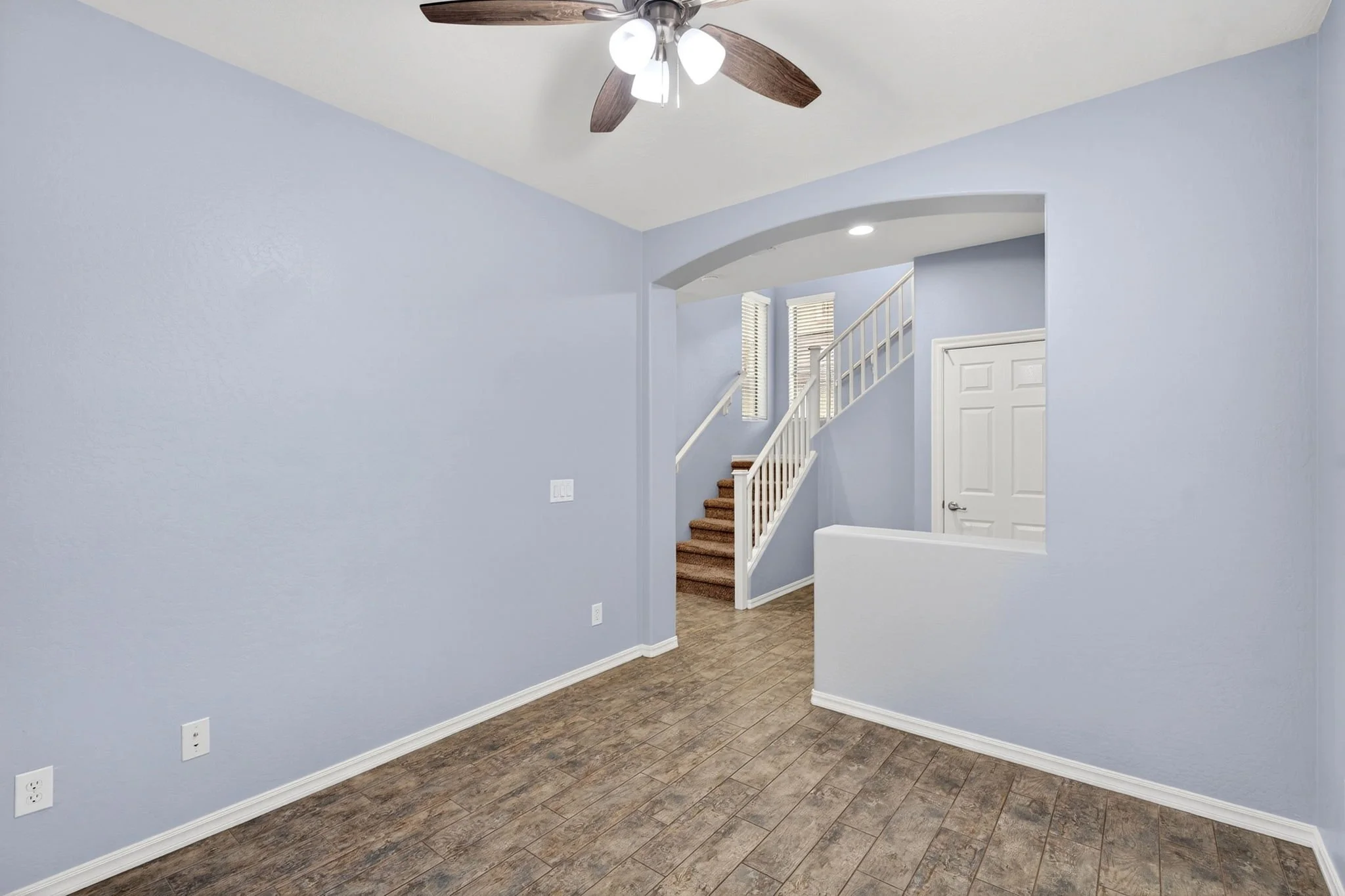 Interior of a house with light blue walls, wooden flooring, a staircase with a white railing, a white door, and a ceiling fan with wooden blades and four lights.