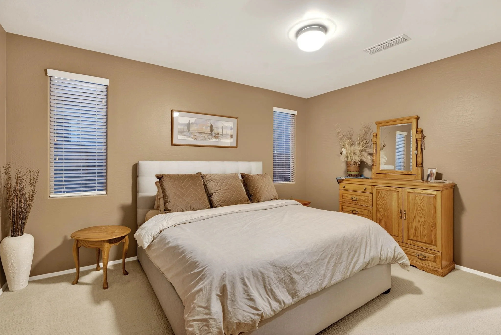 A cozy bedroom with a white bed, brown pillows, wooden nightstand, dresser with mirror, and two windows with blinds, beige walls, and a ceiling light.