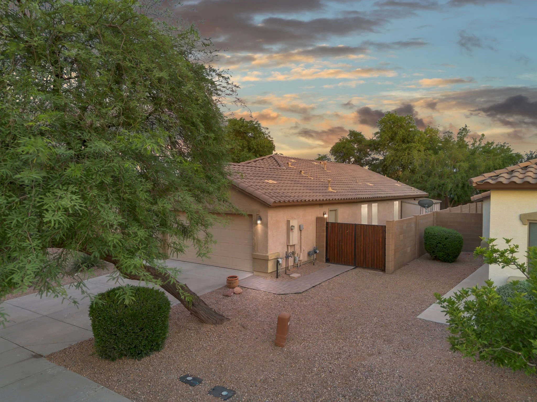 Residential backyard with gravel and concrete pathways, manicured bushes, large trees, a two-story house with a tiled roof, a gate, and an evening sky with clouds.