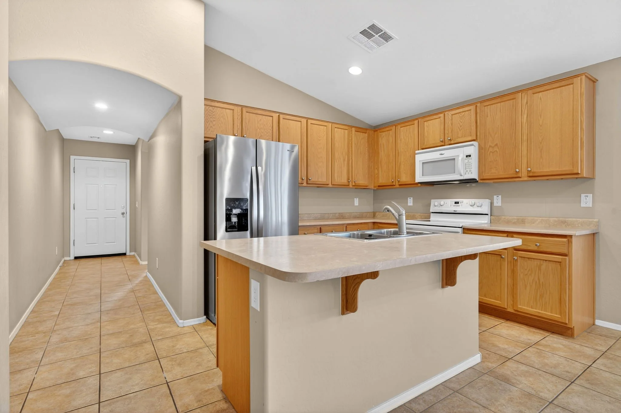 Kitchen area with wooden cabinets, white microwave, stainless steel refrigerator, beige countertops, and tiled floor.