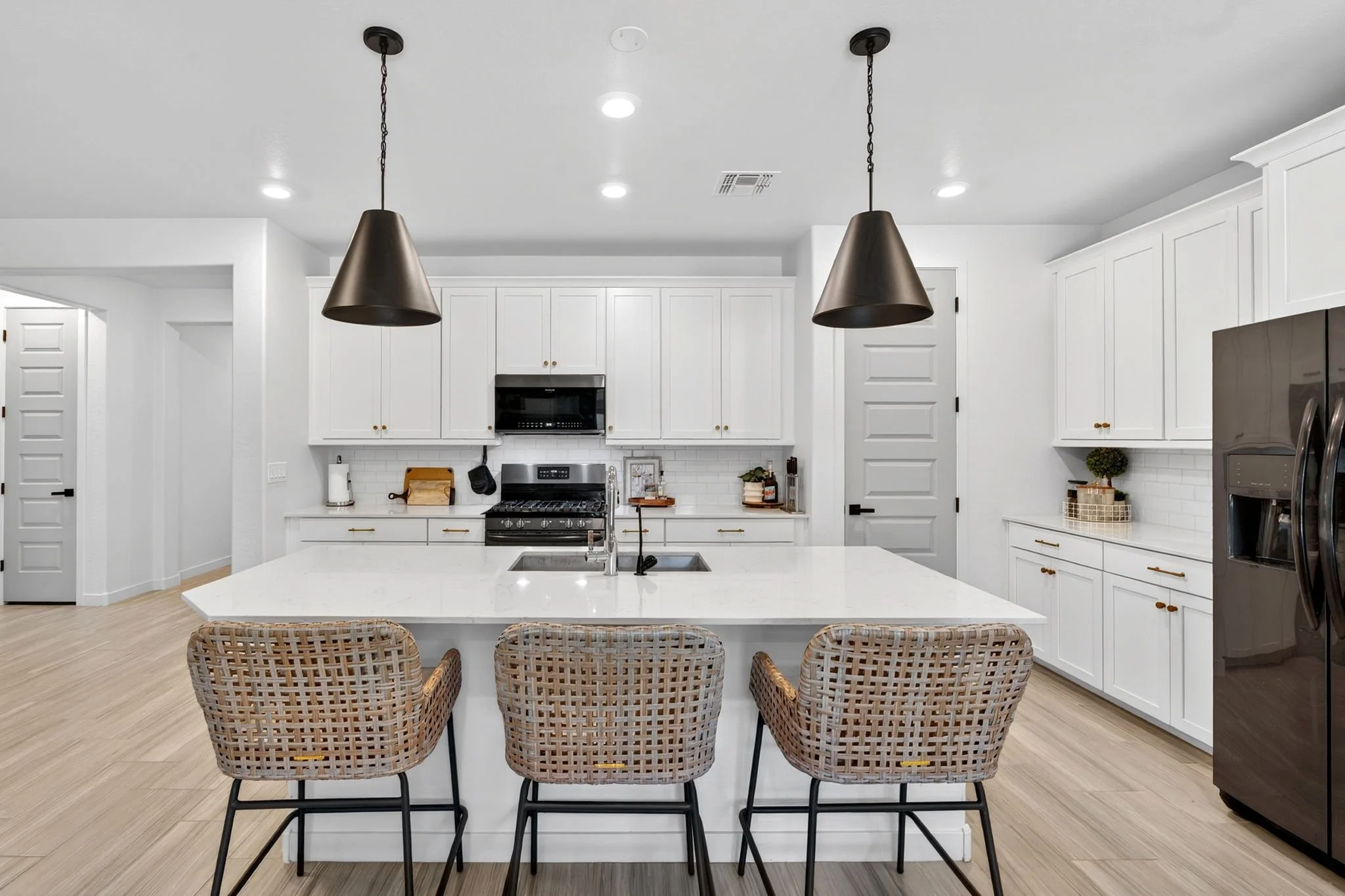 Modern kitchen with white cabinets, a large island with a white countertop, three woven wicker chairs, black pendant lights, stainless steel appliances, and light wood flooring.