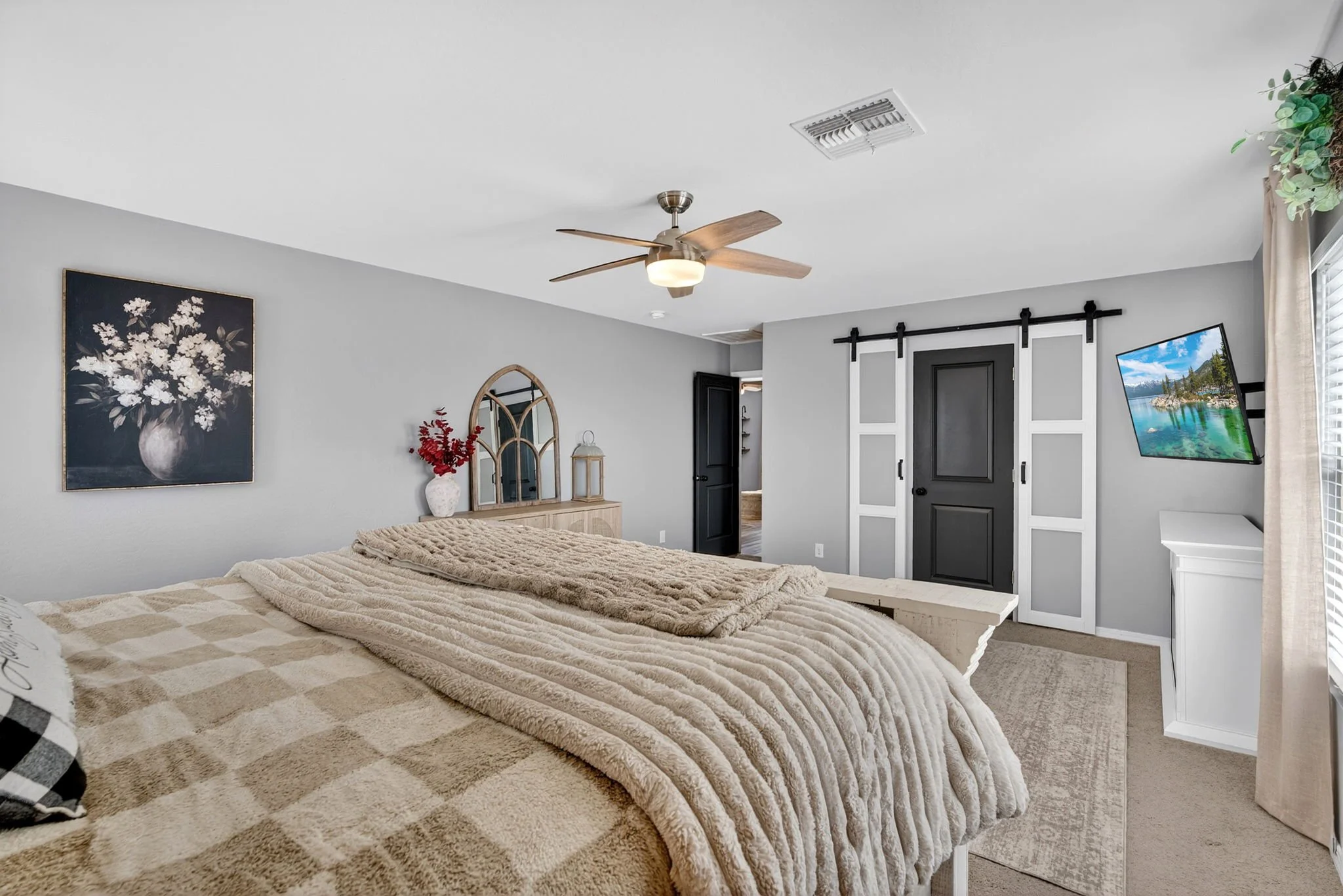 A cozy bedroom featuring a bed with a beige plaid blanket and multiple pillows, a wooden dresser topped with a vase of red flowers, framed floral artwork, a ceiling fan, and a wall-mounted TV near a window with light-colored curtains.