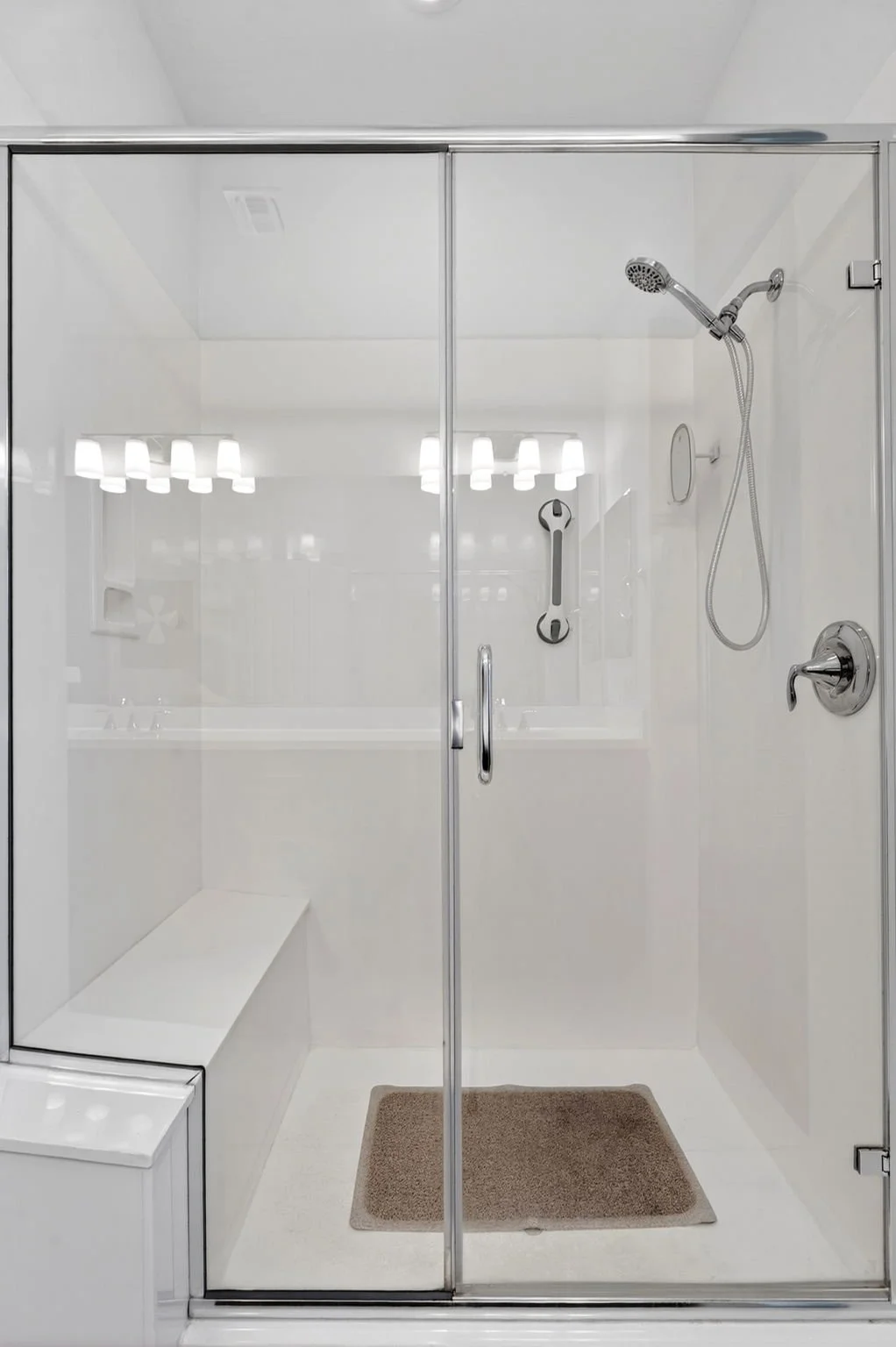 Enclosed white shower with glass door, handheld showerhead, grab bar, small mirror, and a beige bath mat inside.