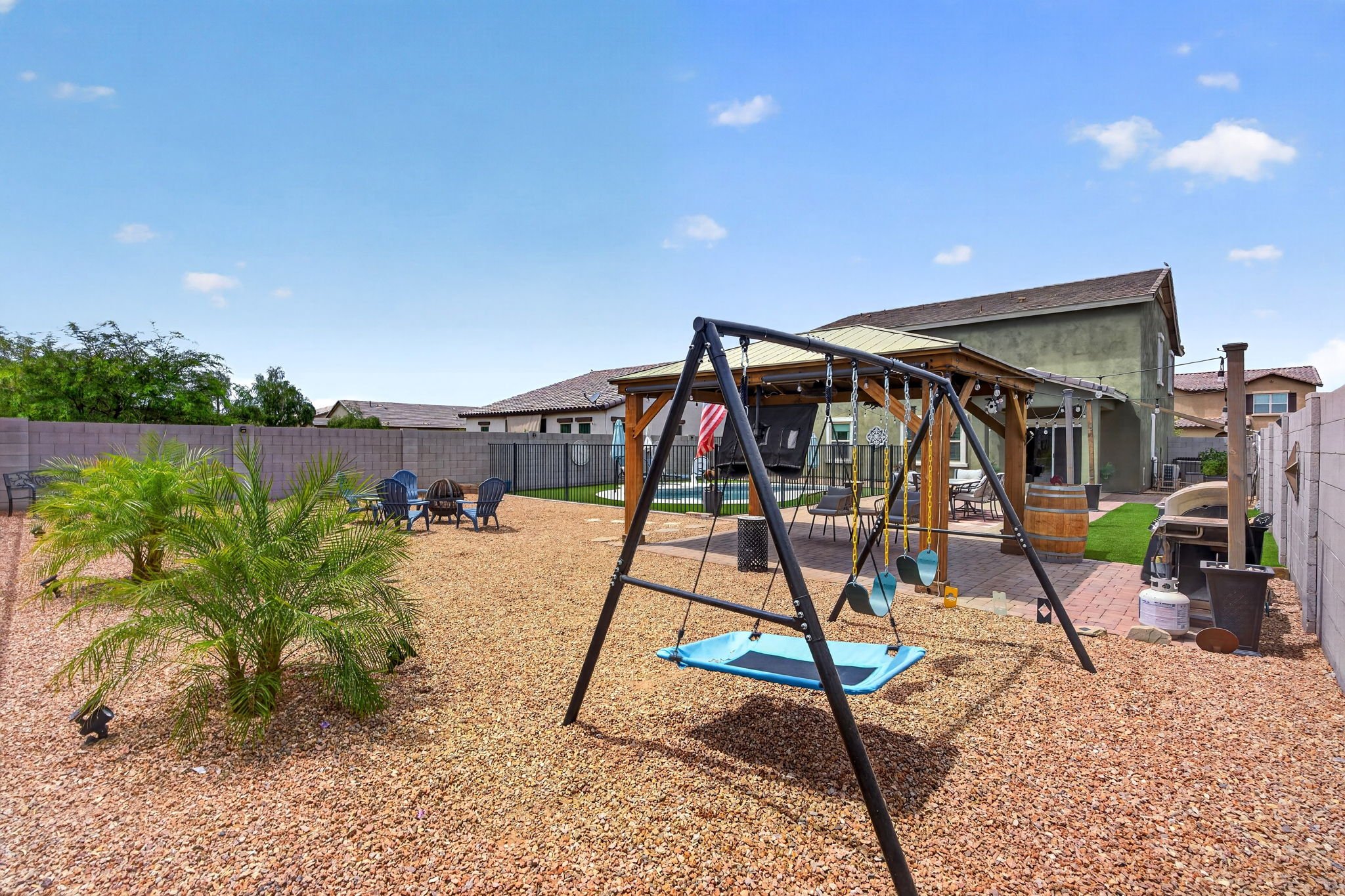 Backyard with a playground swing set, seating area, and small pool, surrounded by a fence and neighboring houses under a blue sky.