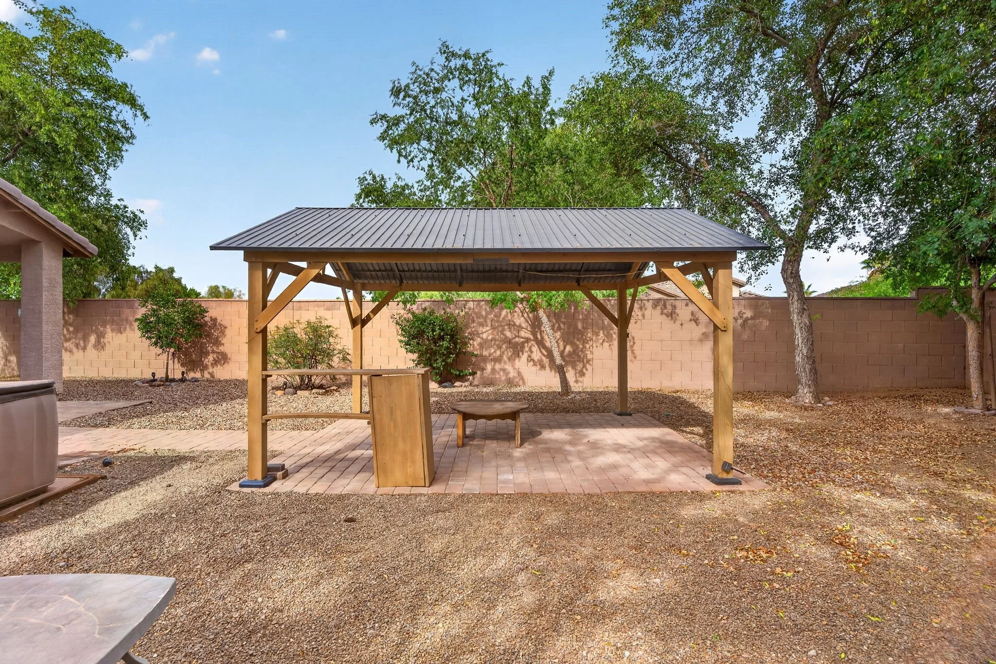 Backyard with a covered wooden shade structure, seating, trees, and a brick patio surrounded by a high brick wall.