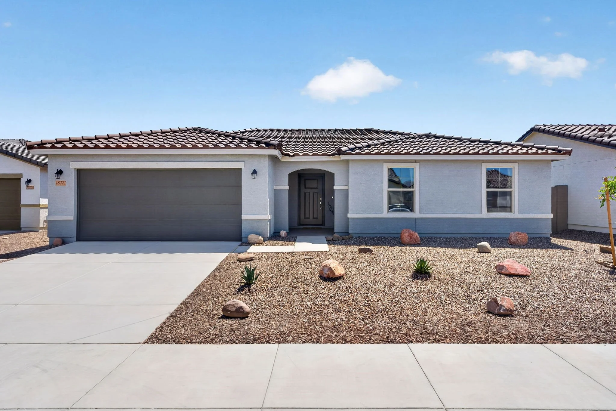 Front view of a modern house with a tiled roof, gray garage door, black front door, and desert landscaping with rocks and small plants under a blue sky.