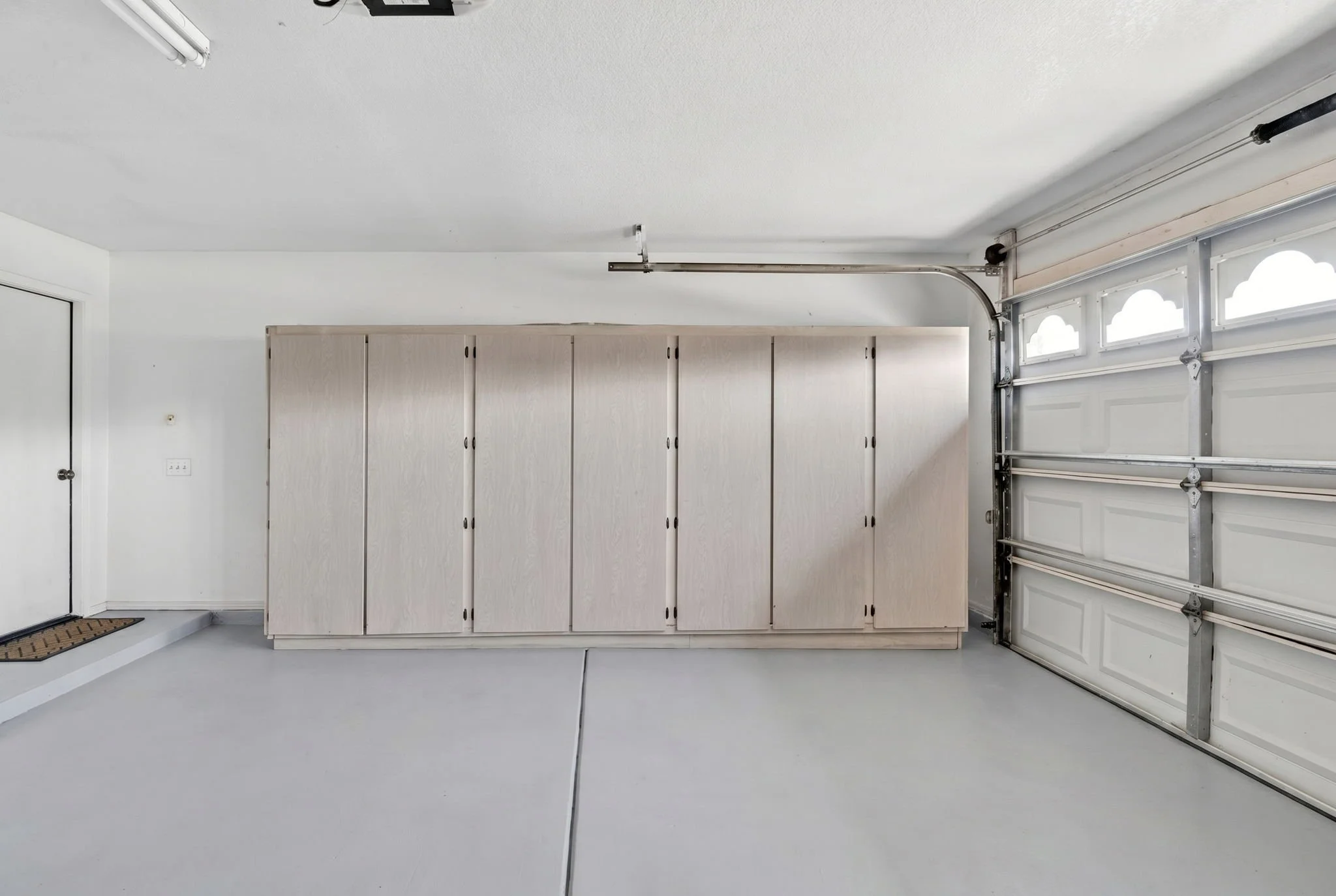 Empty garage with a closed white garage door, beige cabinets along the wall, a door to the left, and a gray floor.
