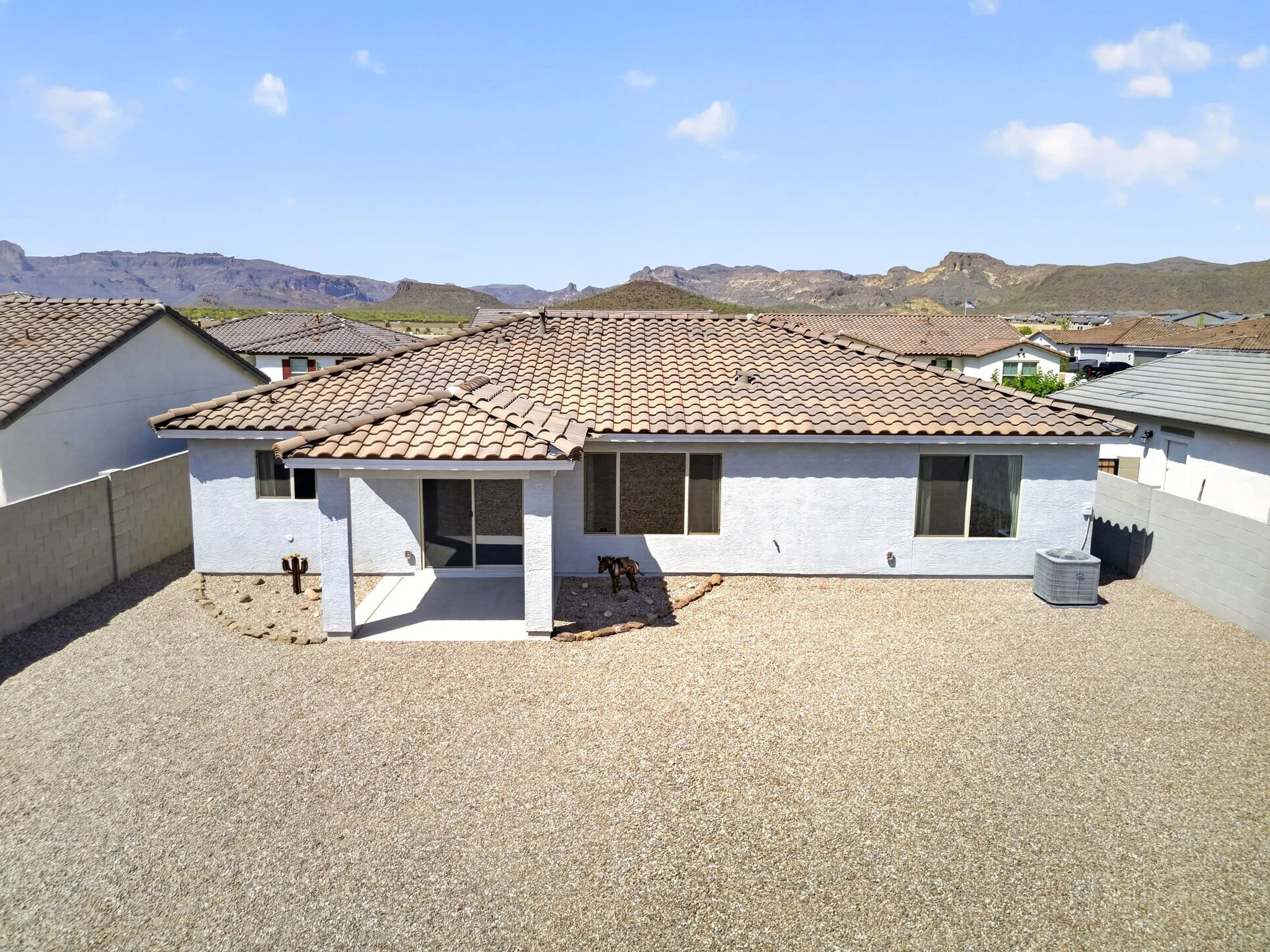 Backyard view of a house with a gravel yard, white exterior walls, tiled roof, and mountains in the background with a few clouds in the sky.