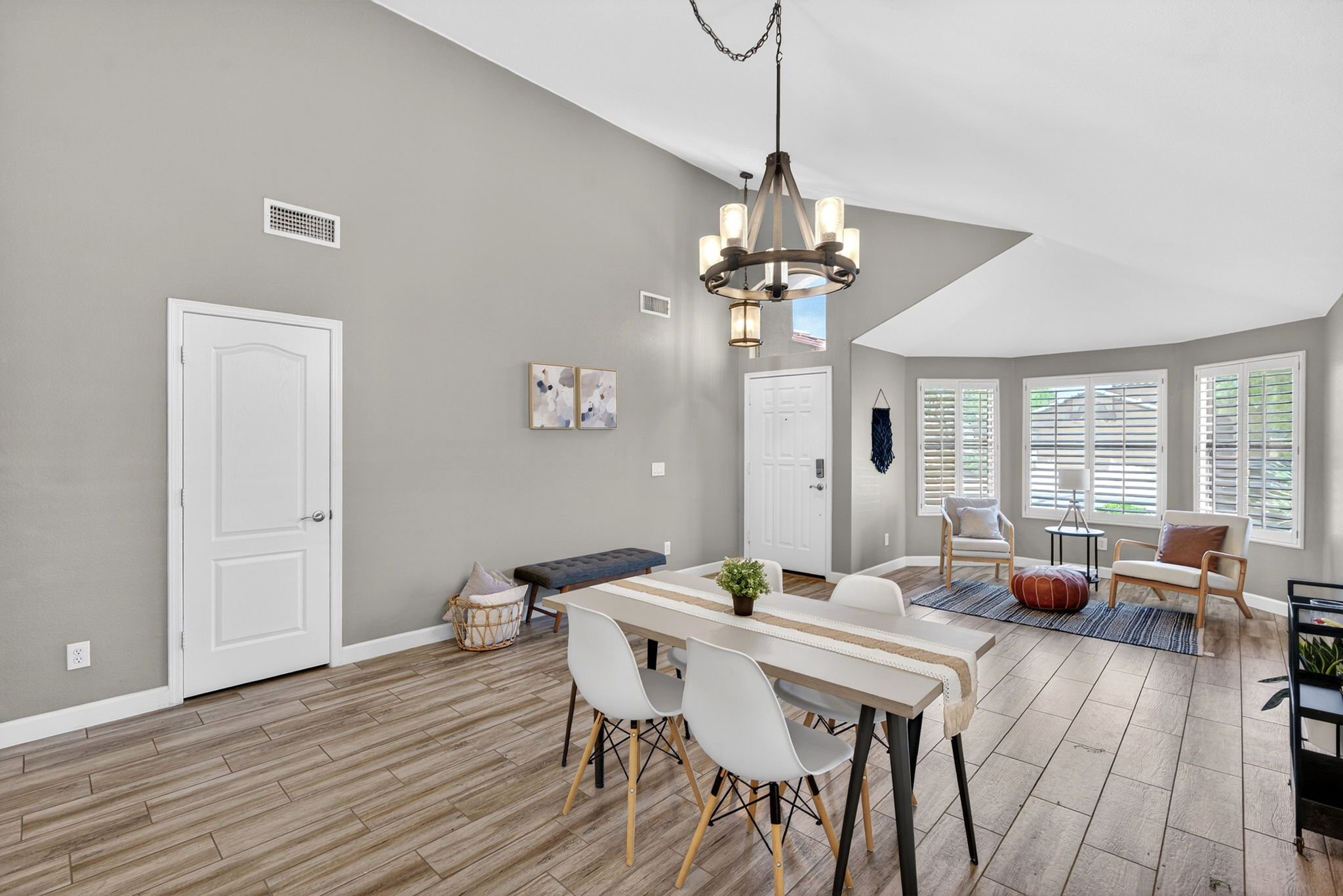Bright living room with light gray walls, large windows with white blinds, modern chandelier, and wooden flooring. Contains a dining table with white chairs and cozy seating area with armchairs, ottoman, and a small side table.