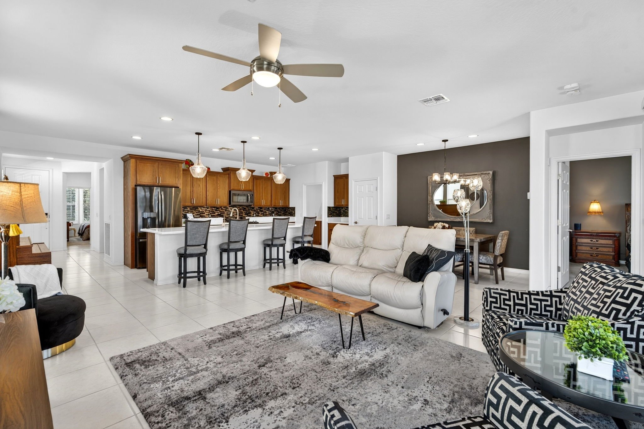 Open-concept living room and kitchen with white tile flooring, white walls, wooden kitchen cabinets, a gray accent wall, and ceiling lights. The living area has a white couch, a black and white patterned chair, and a glass coffee table. Pendant light