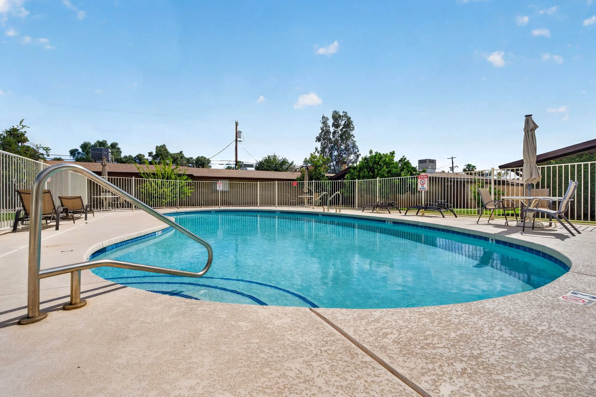 A swimming pool area on a sunny day with chairs, umbrellas, and a fence, with rooftops, trees, and a blue sky with clouds in the background.