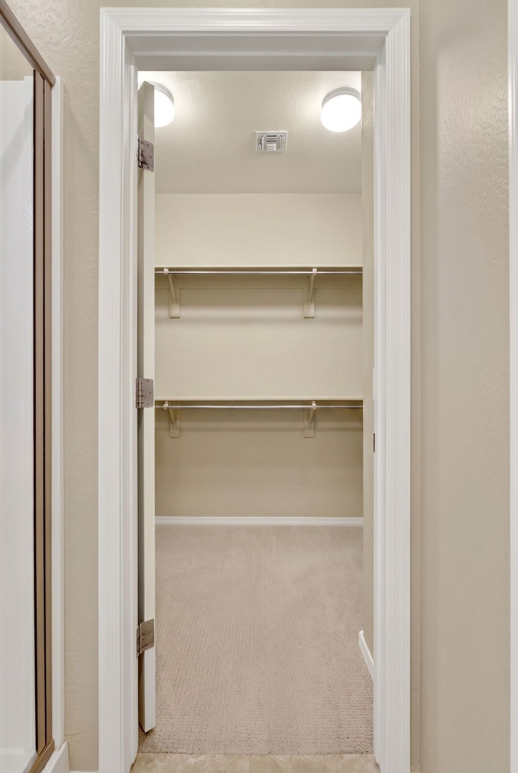 Empty walk-in closet with beige carpet, two hanging rods, and a shelf, illuminated by ceiling lights.