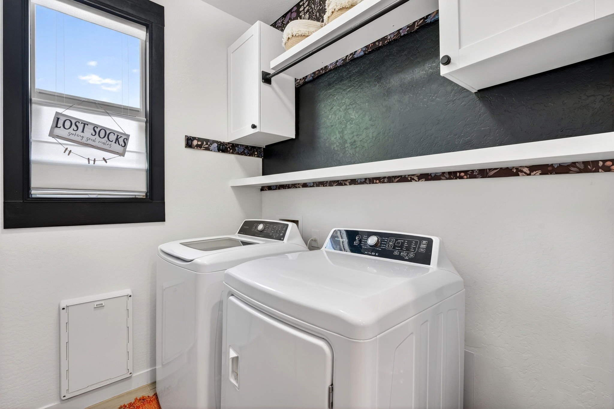 Laundry room with a top-loading washing machine and an adjacent dryer, white walls, a window with a blind, and a small storage cabinet nearby.
