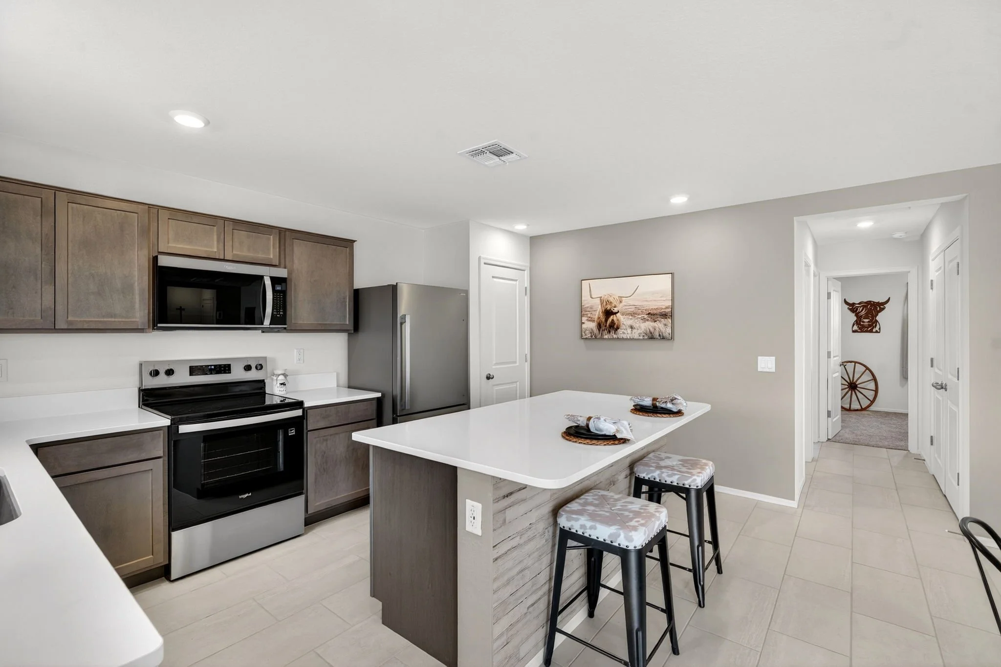A modern kitchen with wooden cabinets, black stove, stainless steel refrigerator, and a kitchen island with three bar stools with patterned cushions. The walls are painted light grey, with a framed picture of a Highland cow and a decorative object re