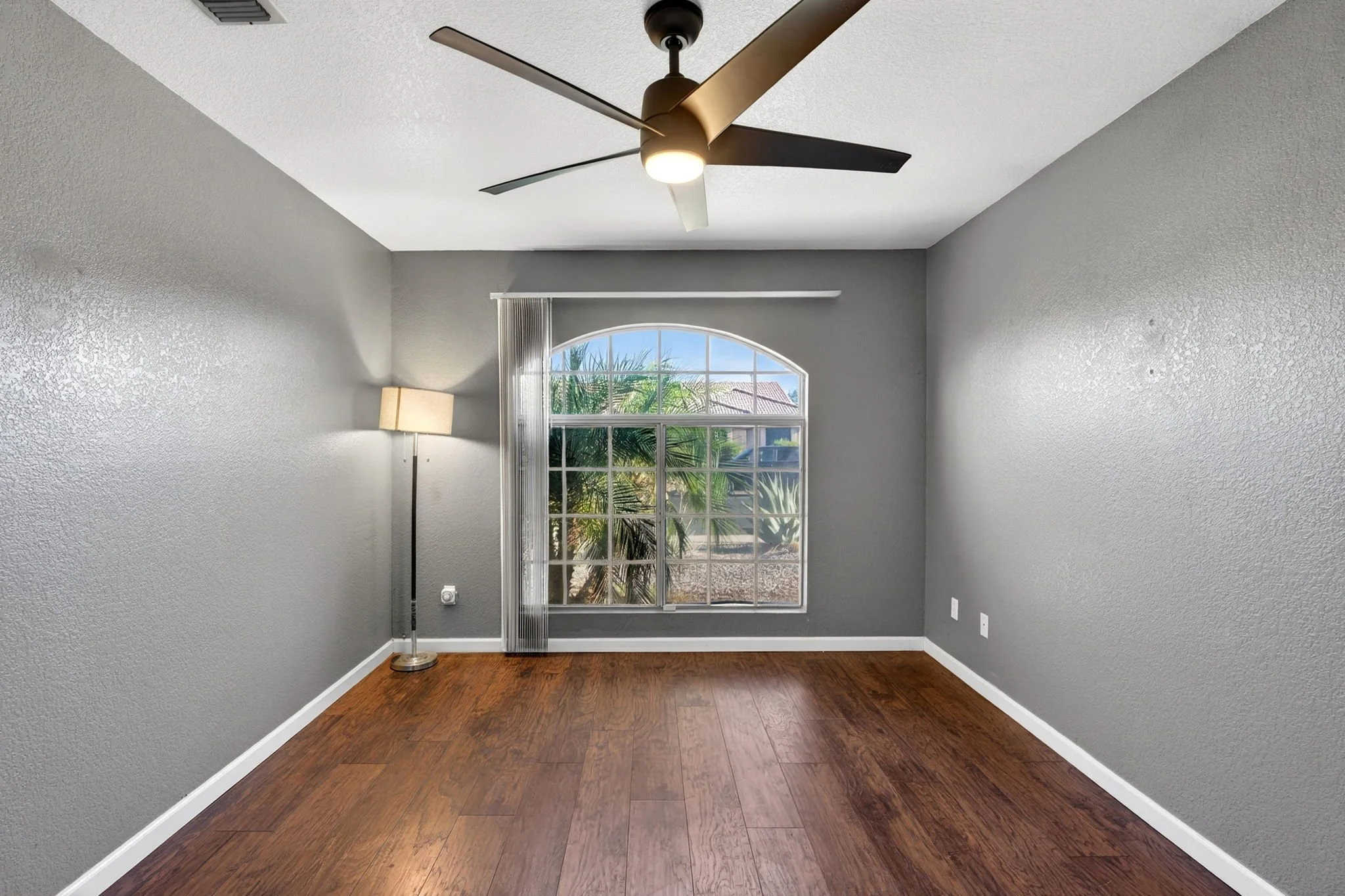 Empty room with gray walls, a ceiling fan, a window with blinds, a floor lamp, and hardwood flooring.