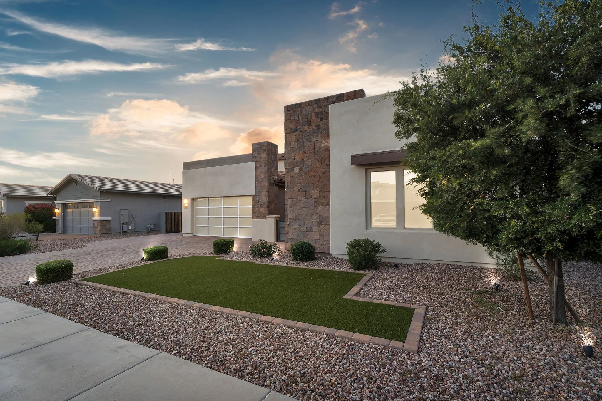 A modern residential house with a landscaped front yard featuring small shrubs, a manicured lawn, and decorative rocks. The house has a white exterior with stone accents and large windows, with a background of a partly cloudy sky during sunset.