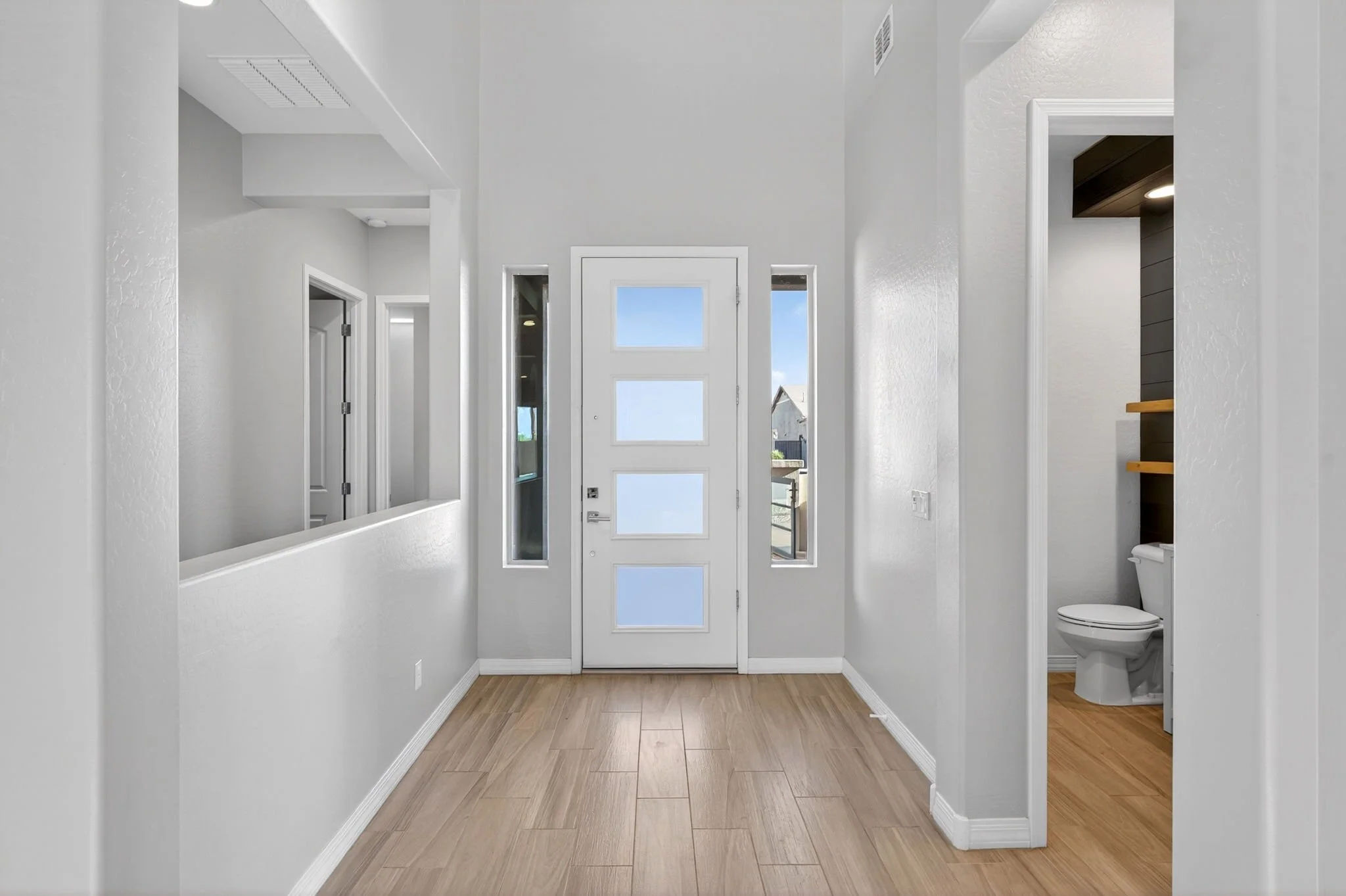Entryway with a white front door featuring four square window panels, flanked by narrow side windows, with a view of neighboring houses outside. The interior has white walls, wood-look tile flooring, and a small bathroom with a toilet visible to the 
