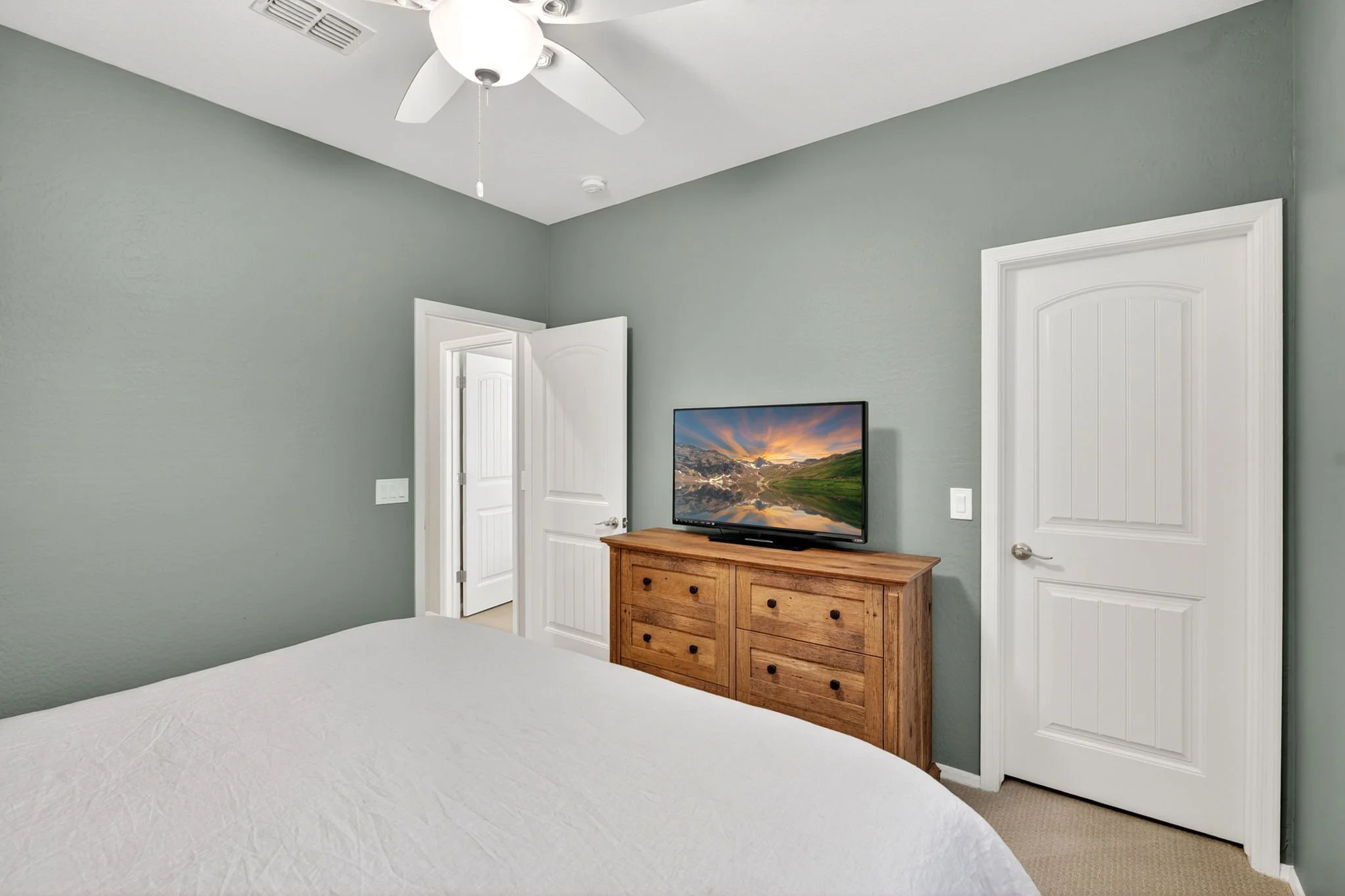 A bedroom with gray walls and white doors, a ceiling fan, and a flat-screen TV on a wooden dresser.