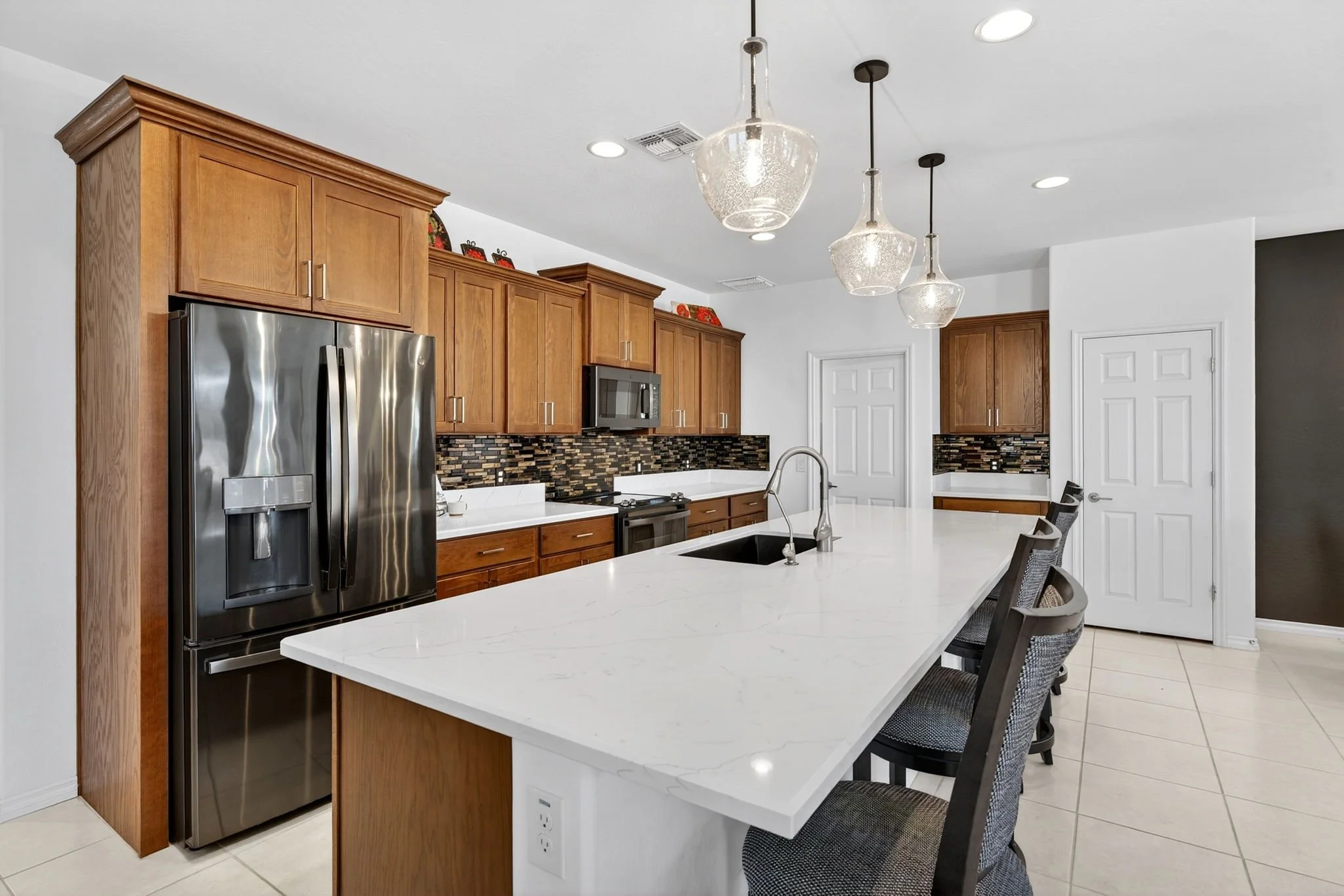 Modern kitchen with wooden cabinets, stainless steel refrigerator, white marble island, pendant lights, and a tiled backsplash.