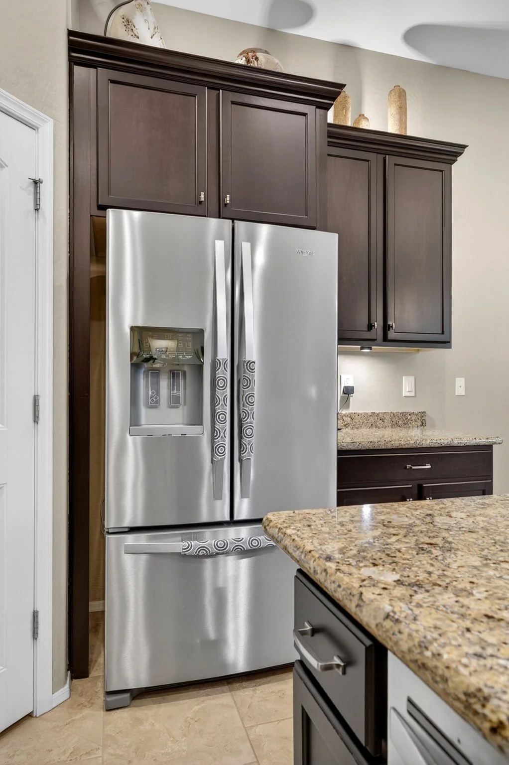 Stainless steel Whirlpool refrigerator with double doors and water/ice dispenser, dark brown wooden kitchen cabinets above, granite countertop, beige wall, and part of a kitchen island.