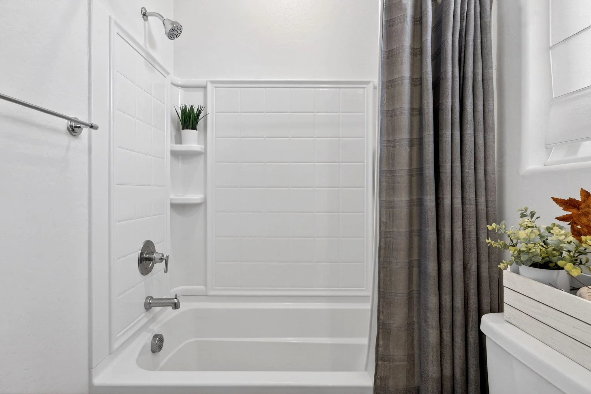 Shower with white tiled walls, a showerhead, and a curtain on the right side. There is a small shelf with a potted plant, and a white wooden stool with artificial plants on it on the right side.